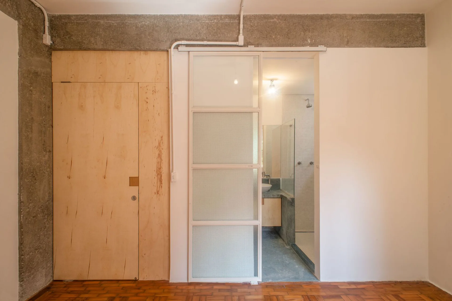 A sliding glass door with a white frame opens to a bathroom with a vessel sink, next to a plywood wall panel and exposed concrete.