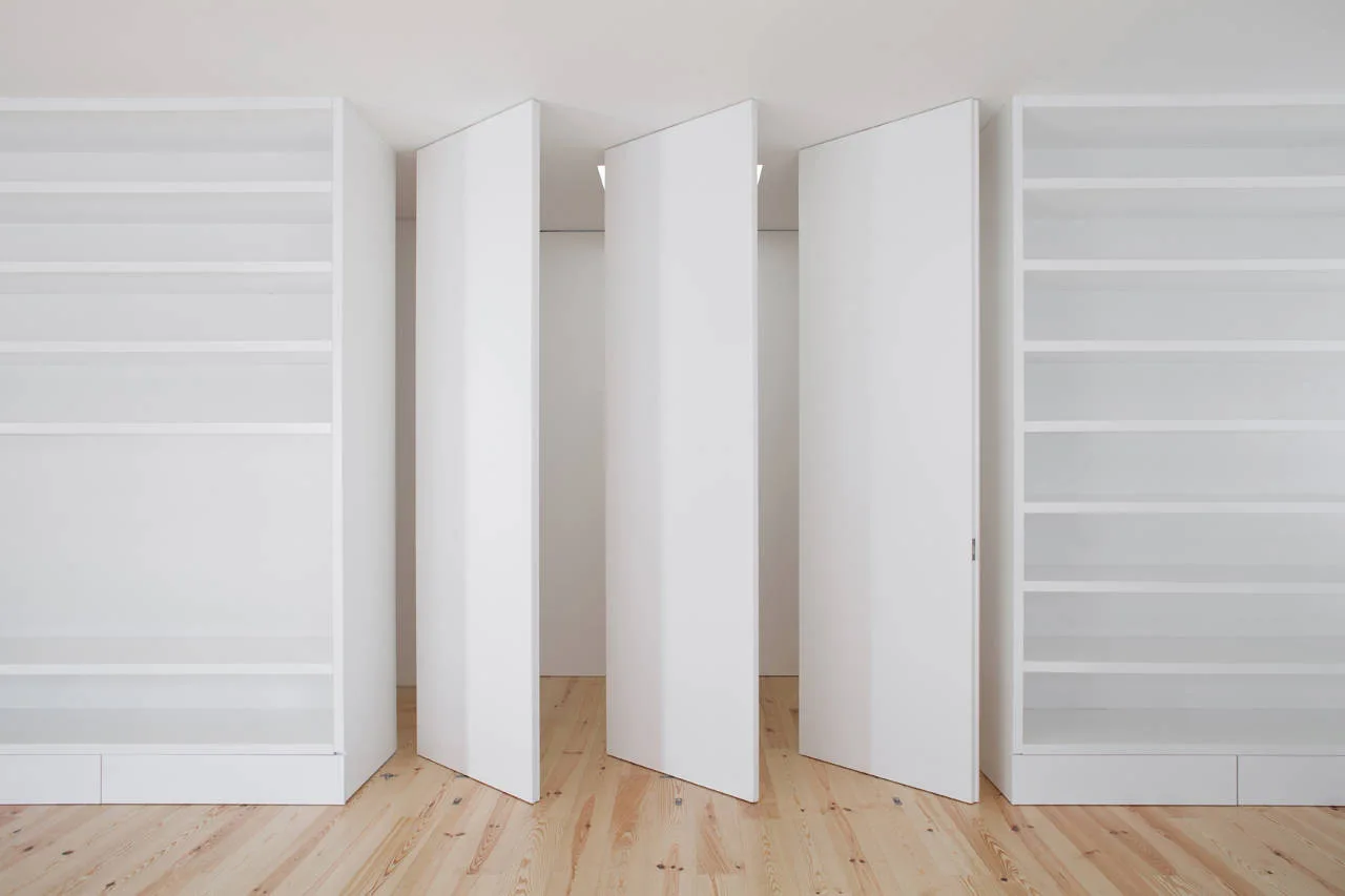 Symmetrical white built-in shelving units flank three floor-to-ceiling pivoting white panel doors on a natural pine wood floor.