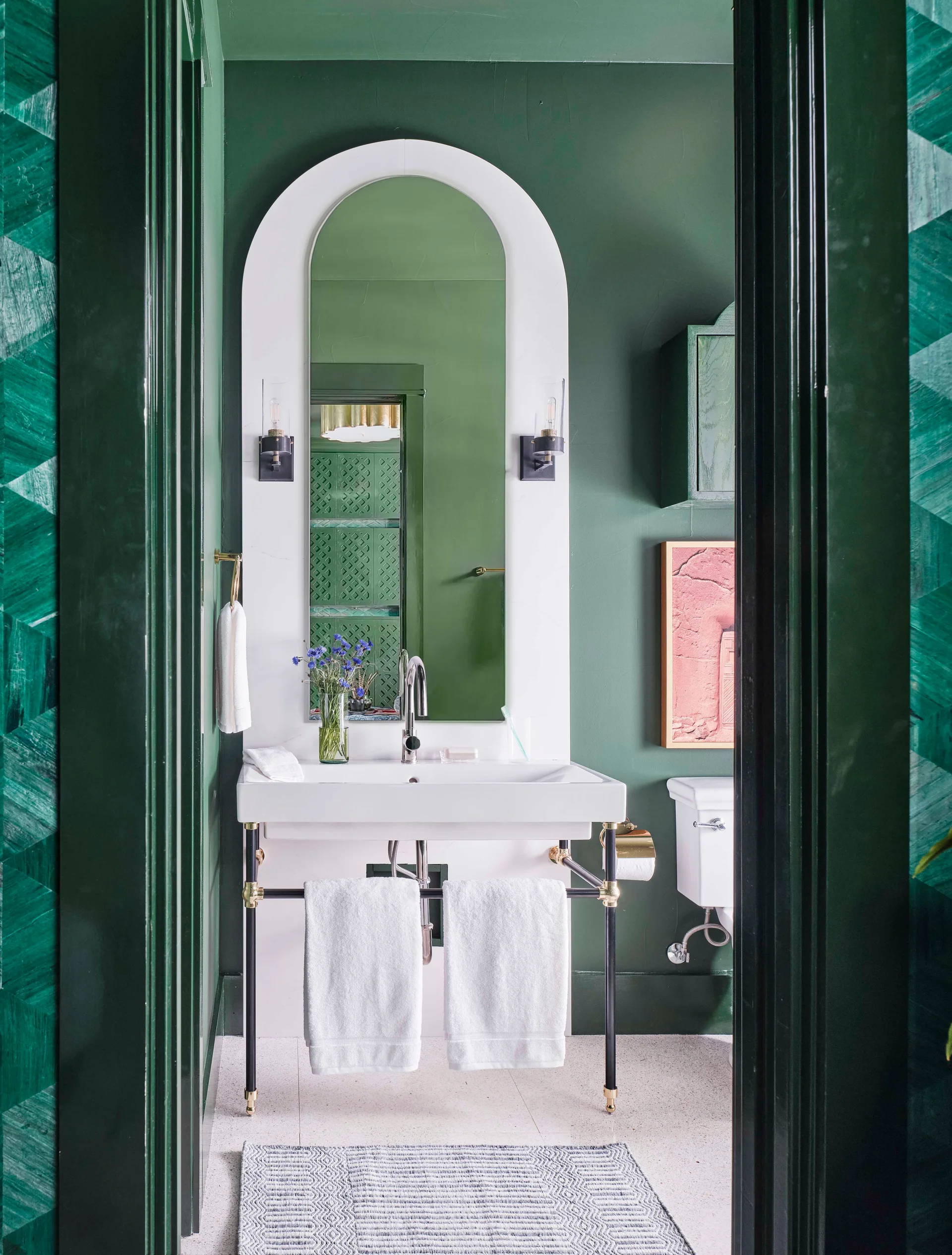 A dark green powder room features a white console sink, an integrated arched mirror, two black sconces, and speckled white flooring.