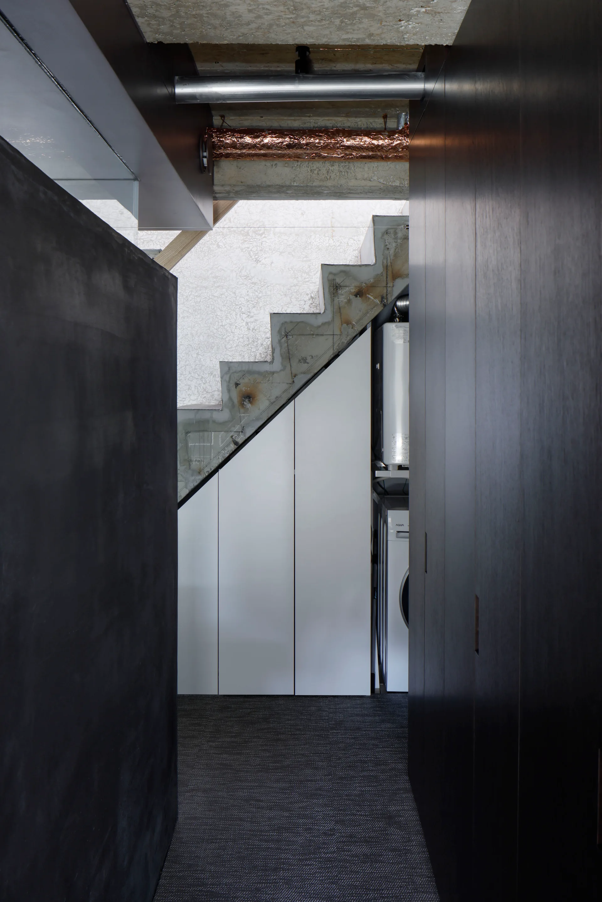 View down a dark hallway showing a raw concrete staircase with integrated white under-stair storage cabinets and exposed ceiling pipes.