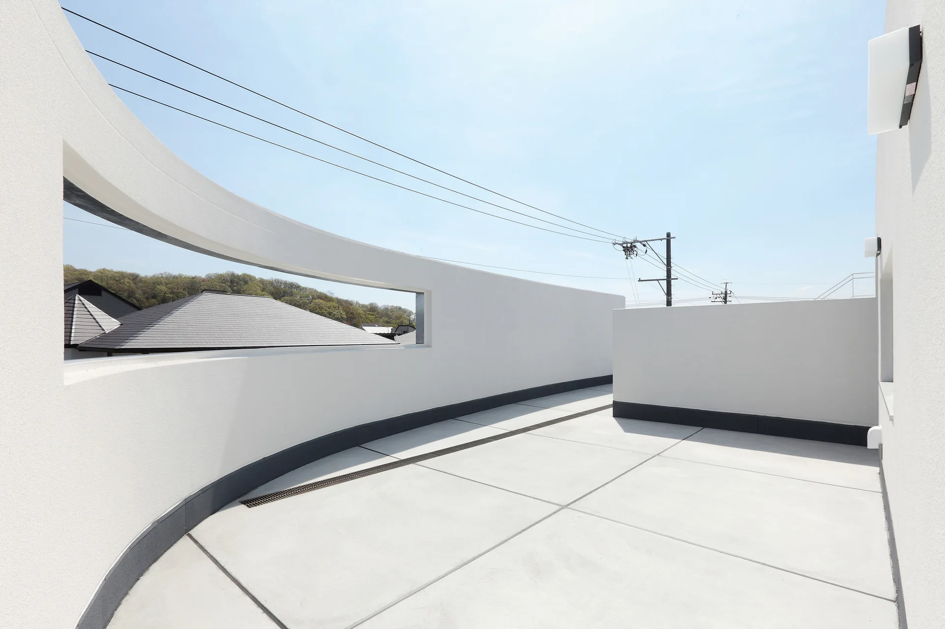 A minimalist rooftop terrace features smooth concrete flooring and a sweeping white curved wall with a rectangular cutout.