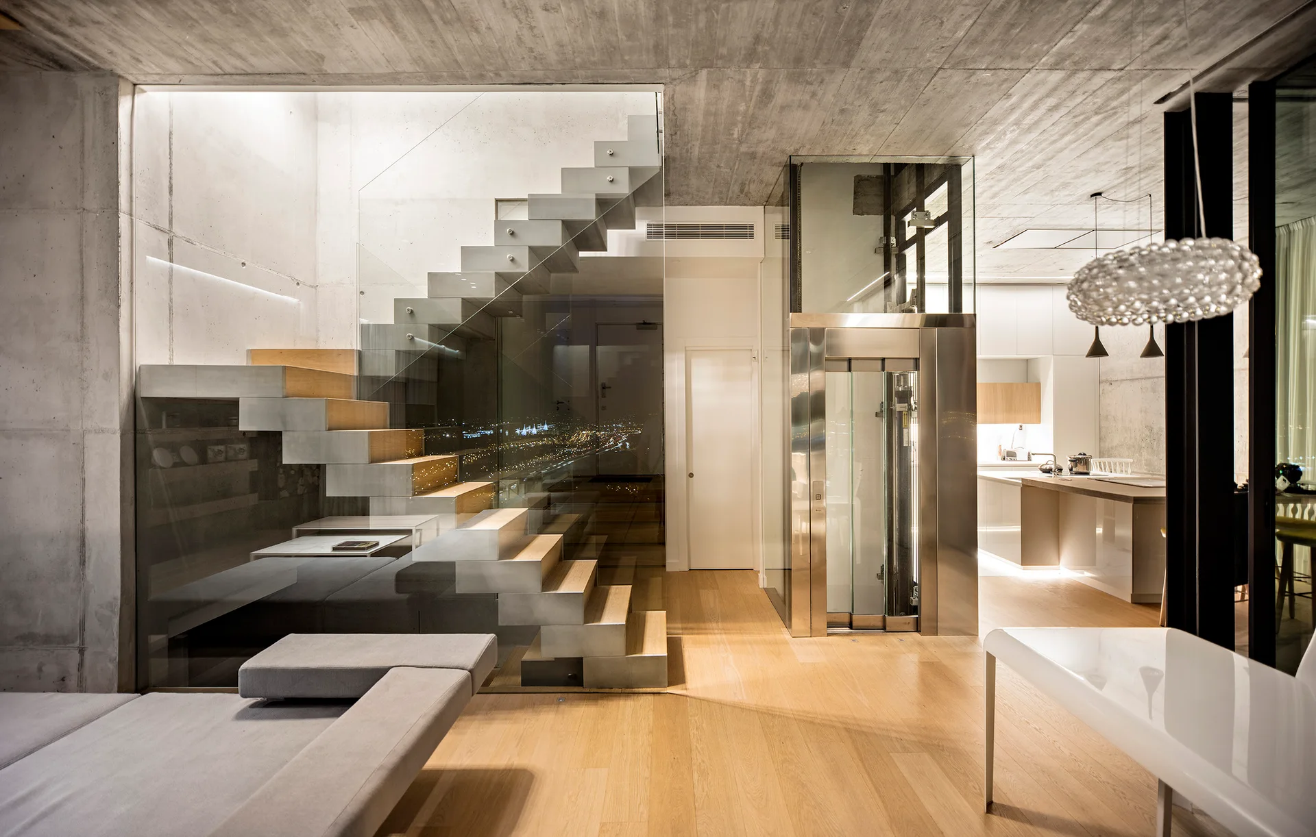 A modern open-plan home featuring a floating staircase with glass walls, a stainless steel residential elevator, and raw concrete ceilings.
