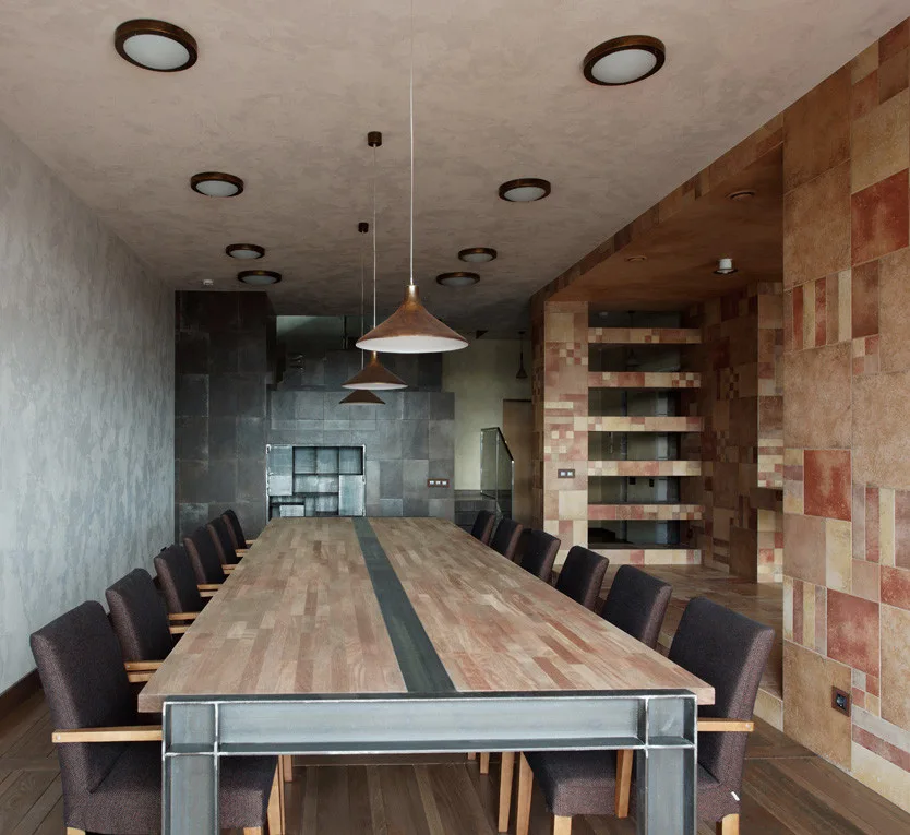 A long wooden dining table with an industrial metal frame is surrounded by dark upholstered chairs beneath three copper pendant lights.