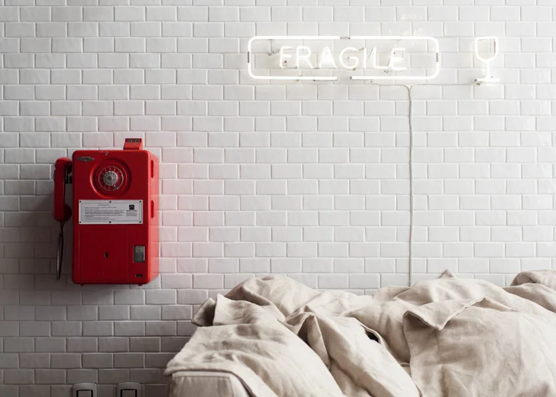 A bright red vintage payphone and a white neon sign hang on a glossy white subway tile wall above a neutral linen sofa.