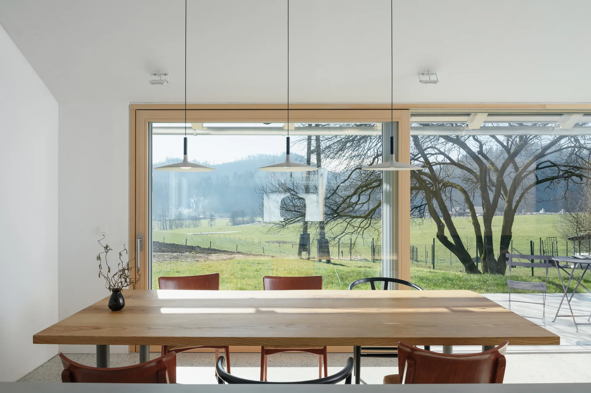 A modern dining room features a long wooden table with various chairs under three white pendant lights, in front of a large picture window.