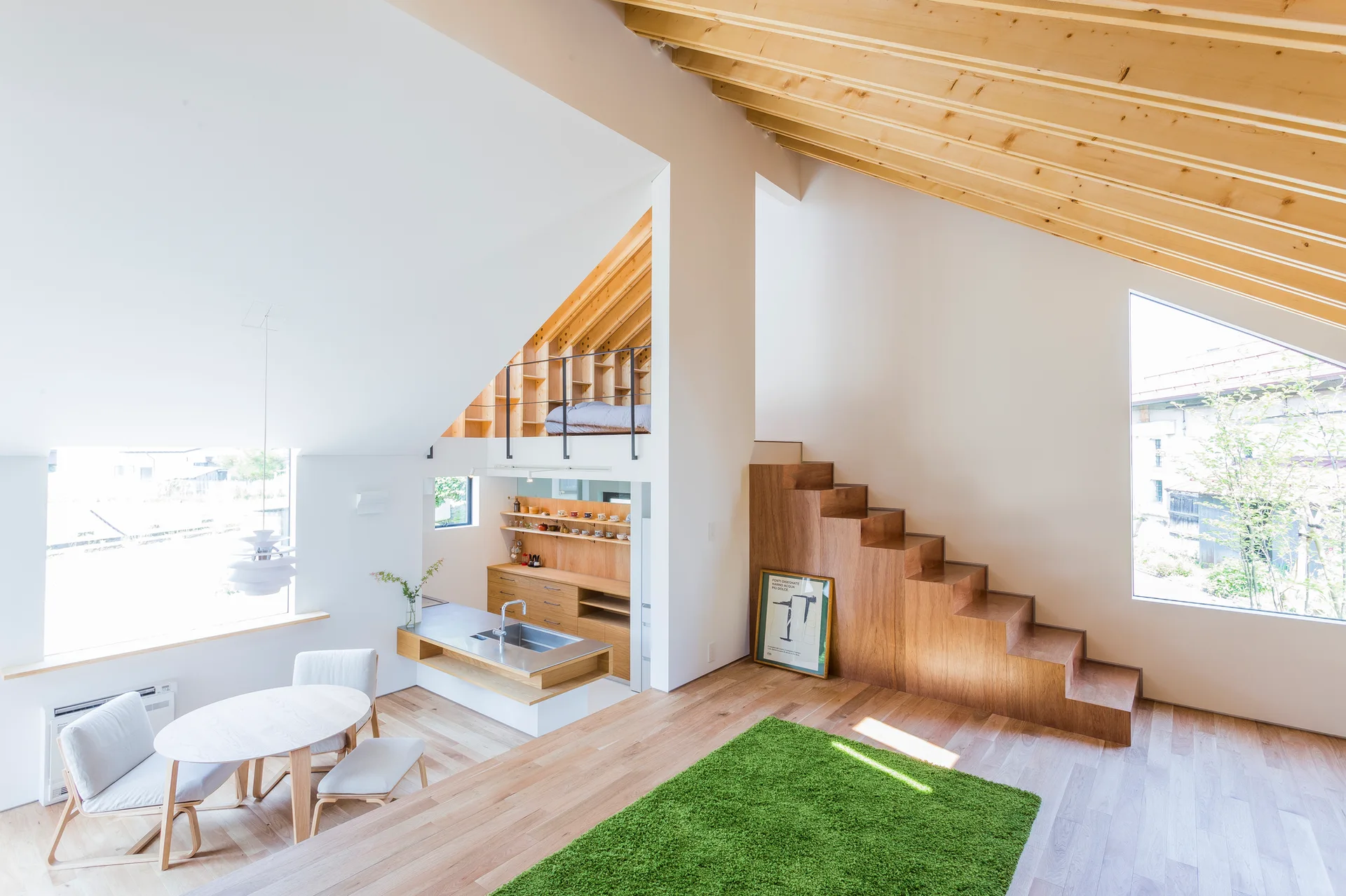 An open-plan living area features a minimalist wooden staircase, a sloped ceiling with exposed beams, a small dining set, and a vibrant green rug.