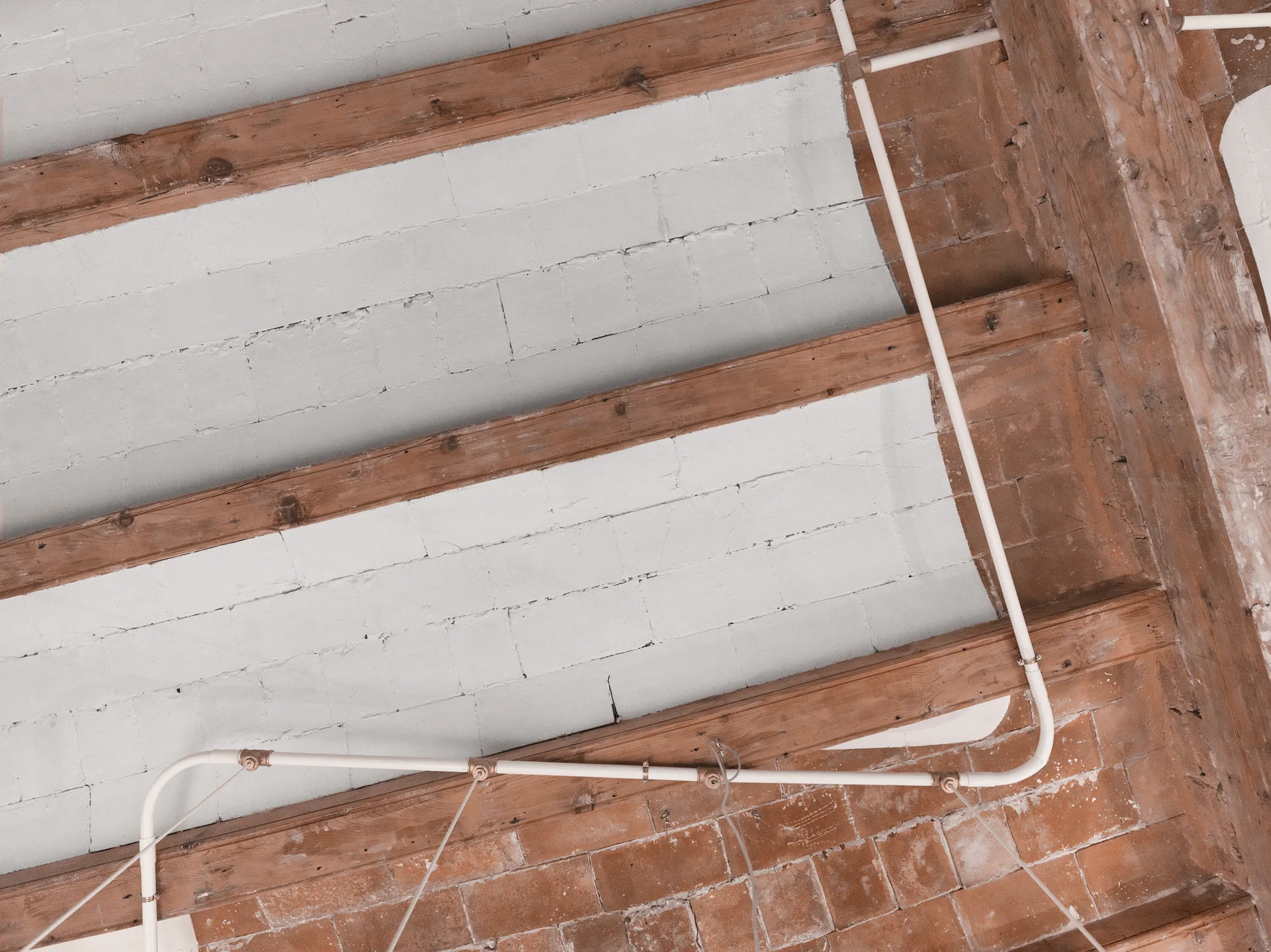 An industrial ceiling features warm wooden beams, white painted masonry, red brick accents, and white electrical conduit pipes.