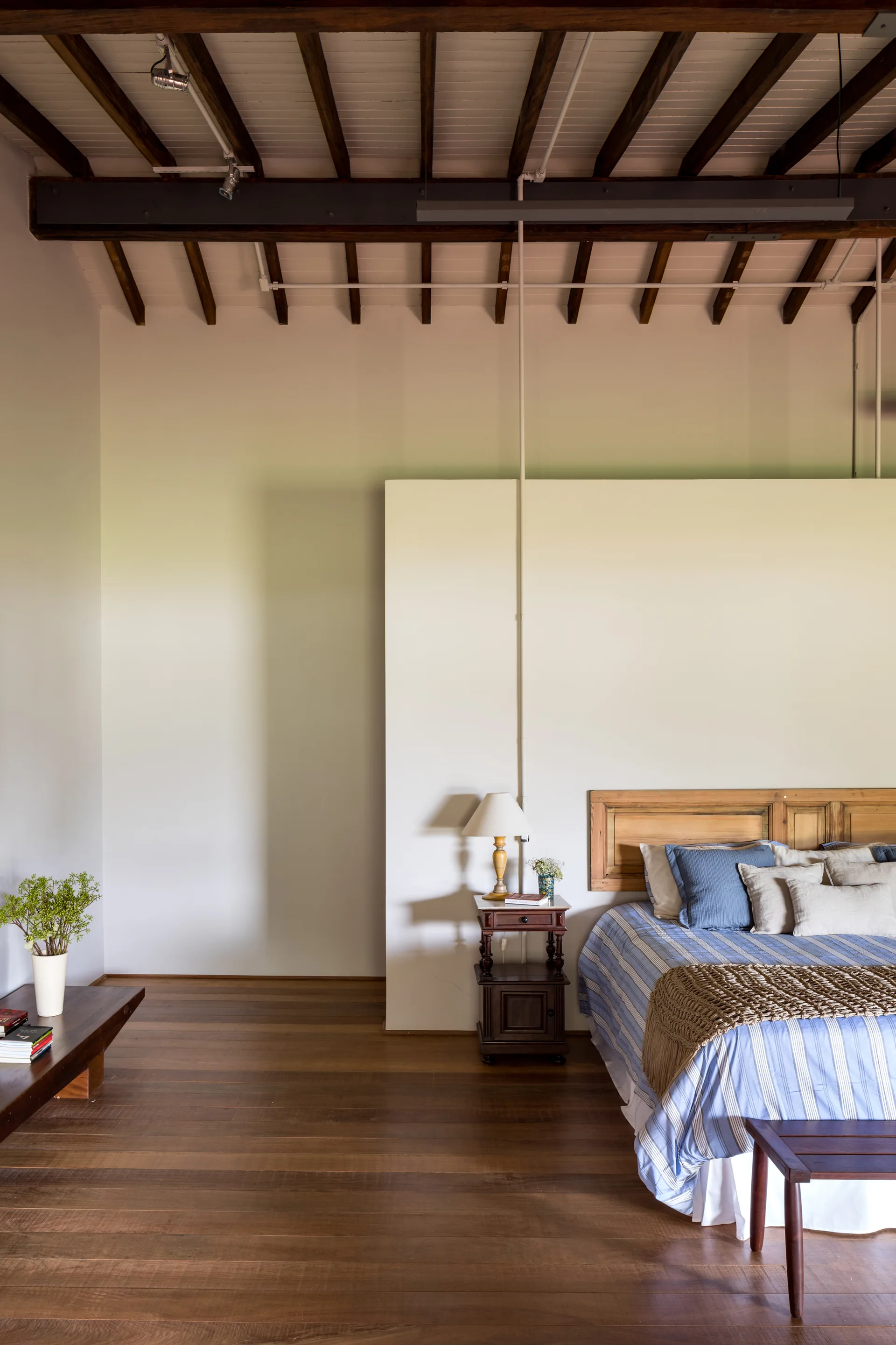 A rustic bedroom features a bed with blue striped bedding next to an off-white partition wall under an exposed dark wood beam ceiling.
