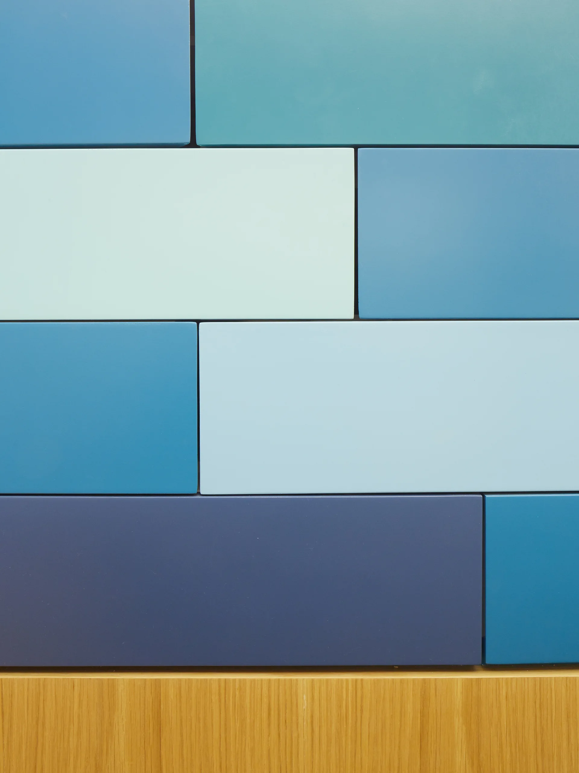 A close-up of rectangular matte tiles in shades of blue and teal arranged in a running bond pattern above a warm oak-toned wood baseboard.