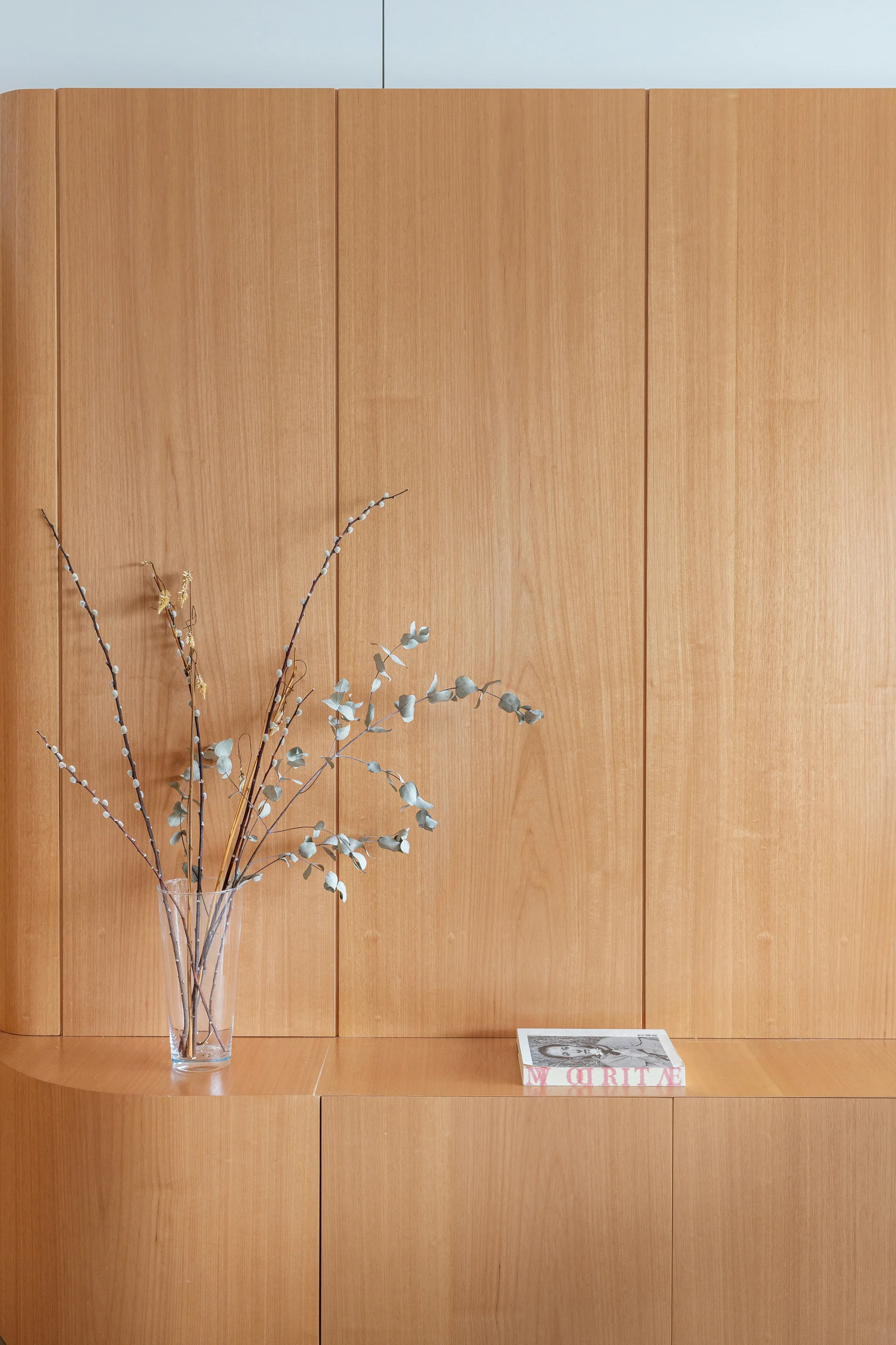 A light wood built-in cabinet with a curved corner sits against matching vertical wall paneling, holding a glass vase with dried branches and a book.