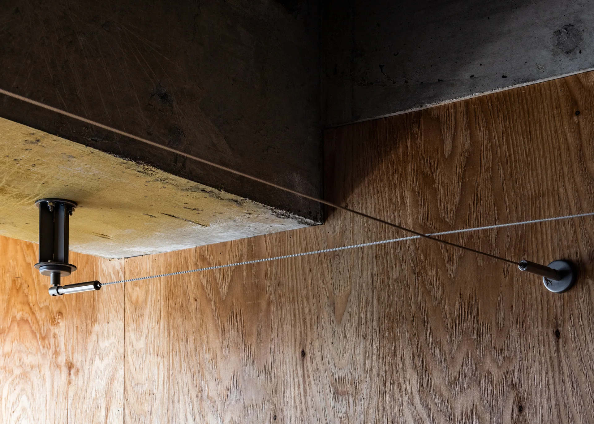 A metal tension cable system is mounted between an exposed concrete ceiling and a plywood paneled wall.