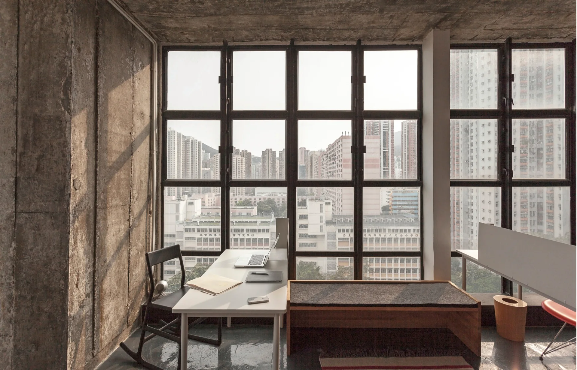 An industrial home office featuring a white desk and a black chair next to a large black grid window and raw concrete walls.