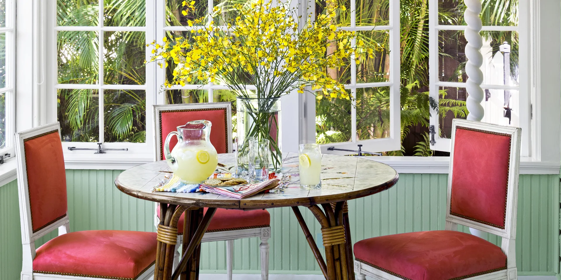 A sunroom dining set features a rattan table and red chairs against mint green beadboard walls and white windows opening to tropical greenery.