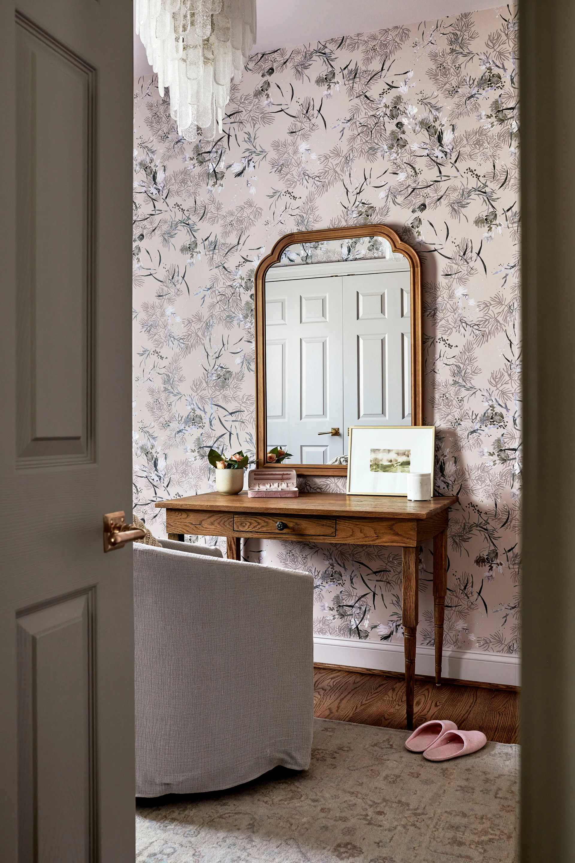 A wooden vanity table with an arched mirror sits against dusty pink botanical wallpaper next to an upholstered armchair and a tiered glass chandelier.