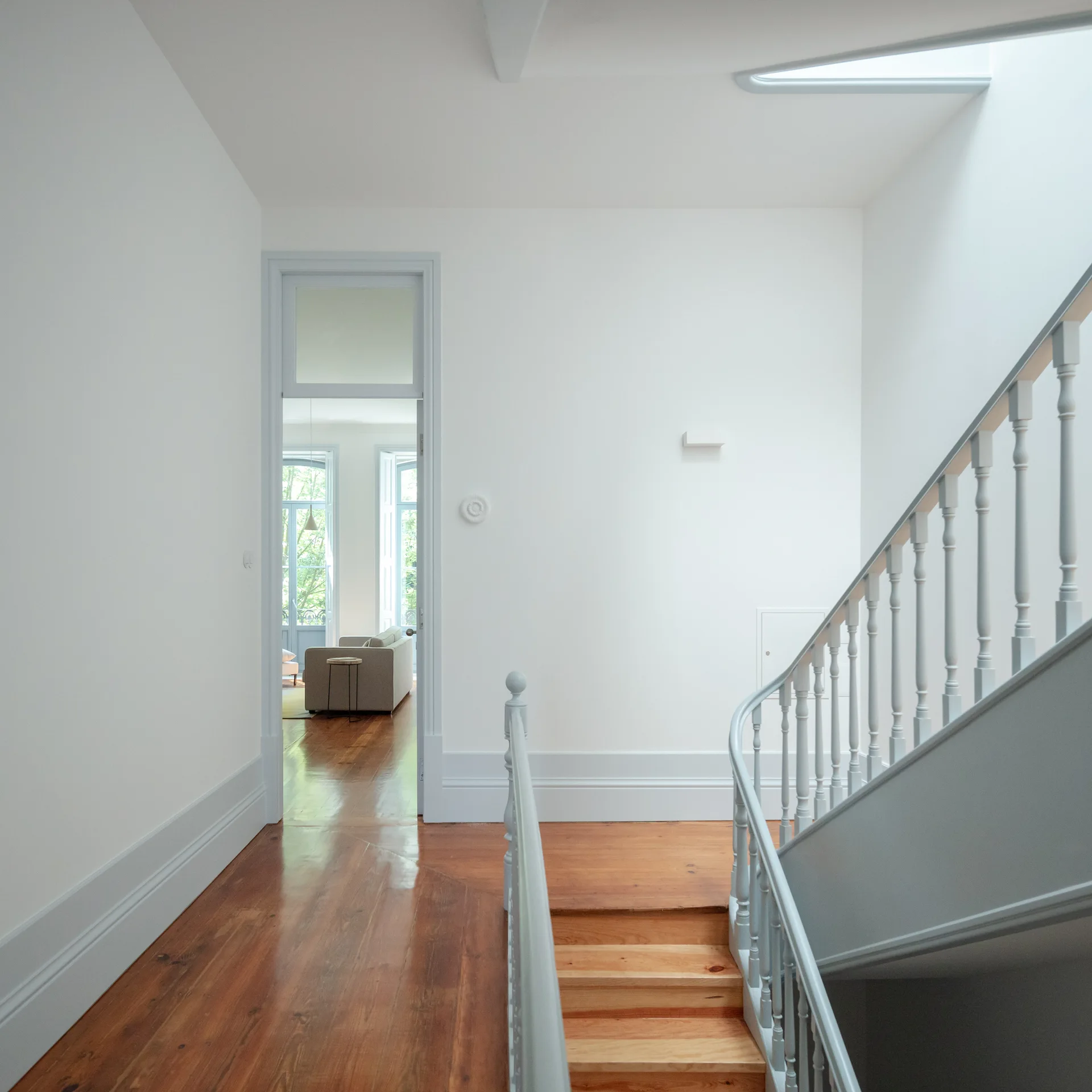 A bright white hallway featuring a wooden staircase with pale blue-grey railings, wood plank floors, and an open doorway leading to a living room.