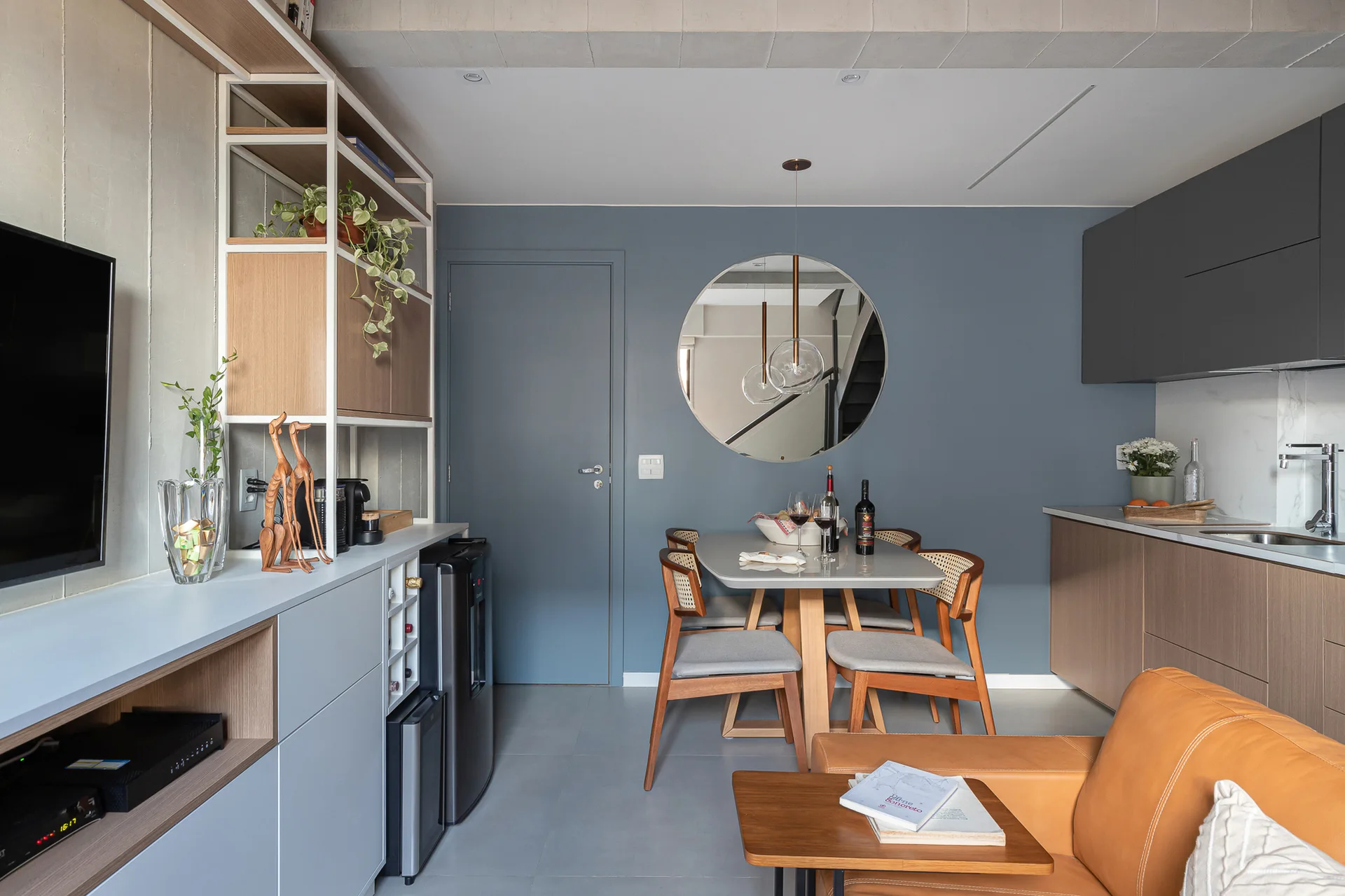 A modern open-plan dining space features a dusty blue wall with a round mirror, cane dining chairs, a grey table, and kitchen cabinets on the right.