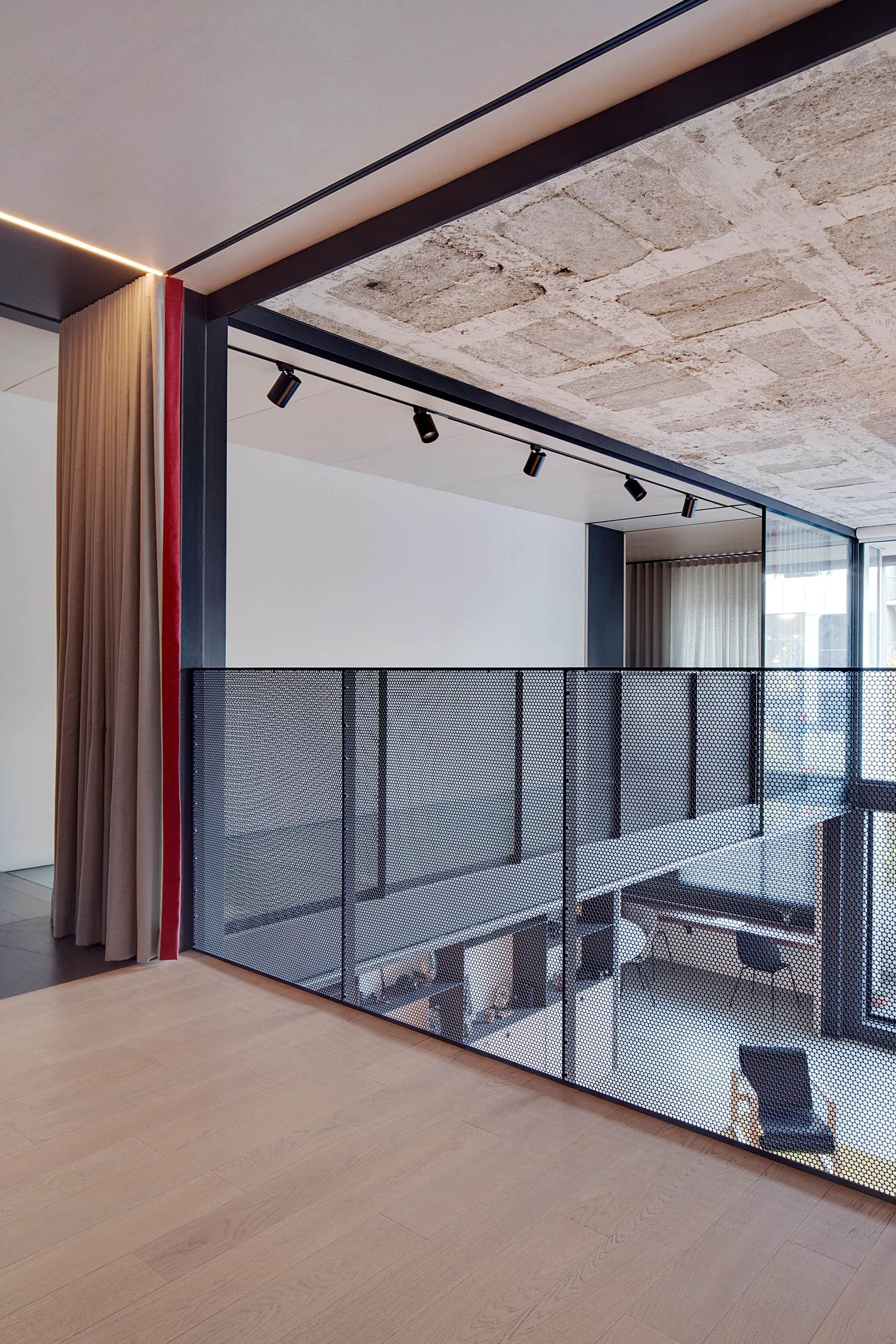 A modern mezzanine landing features light wood flooring, a black perforated mesh railing, track lighting, and a rough concrete ceiling.
