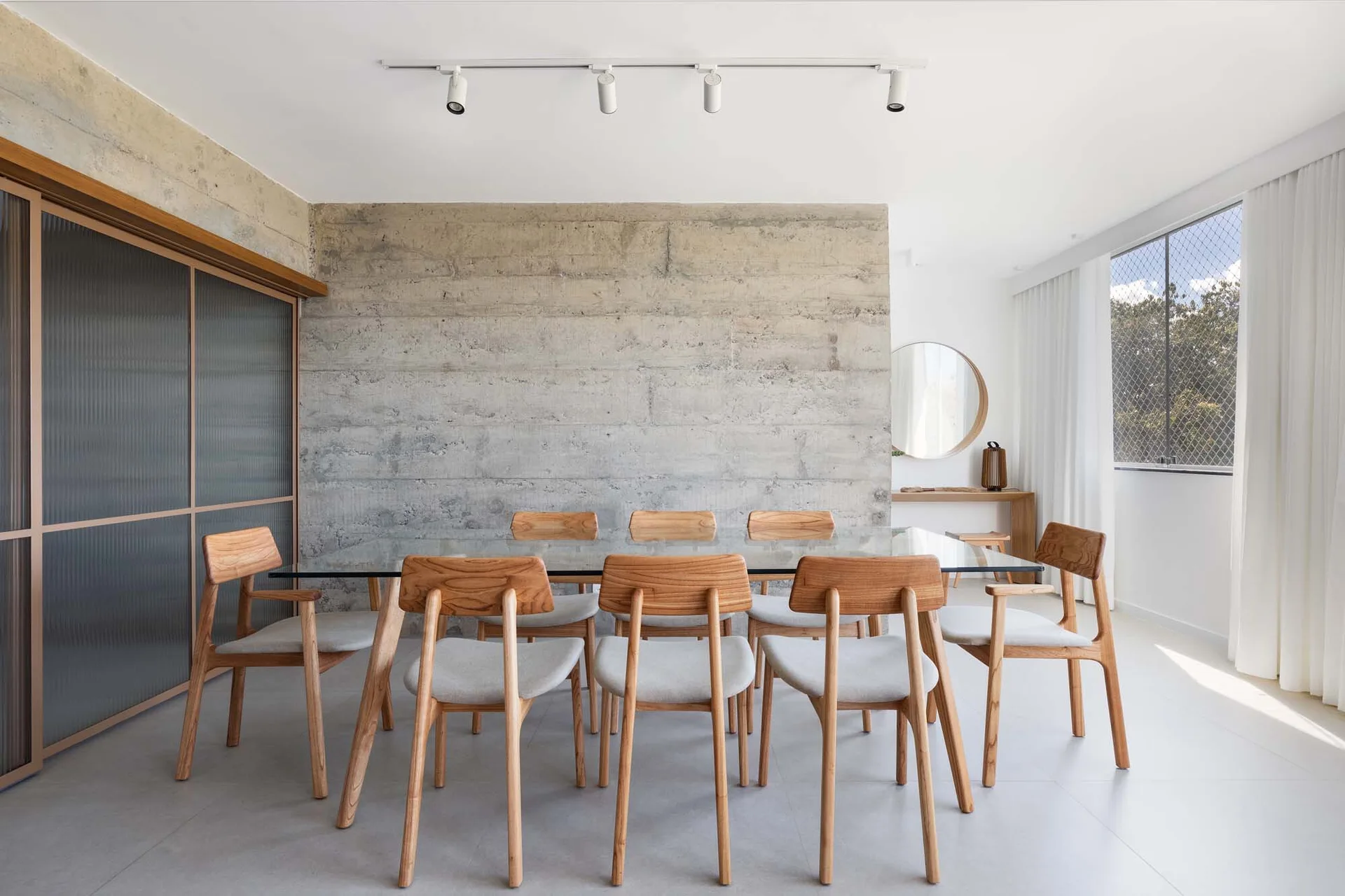 A modern dining room featuring a glass-topped table, wooden chairs with light grey cushions, and a textured raw concrete wall.