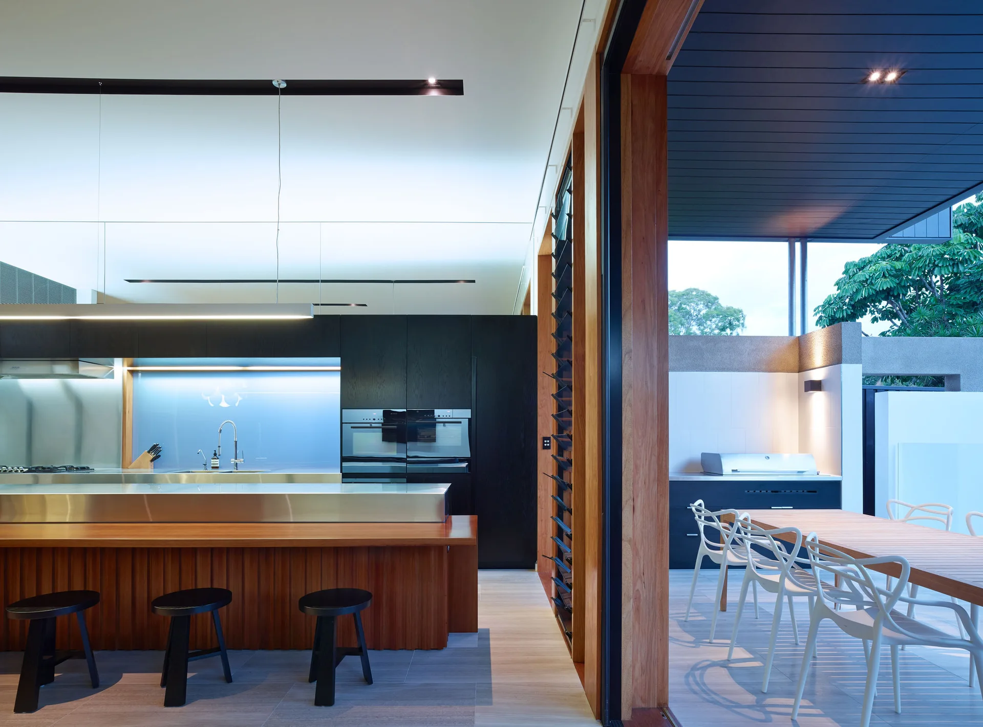 A modern kitchen with a wooden island and steel countertop transitions to an outdoor patio with a wooden dining table and white chairs.