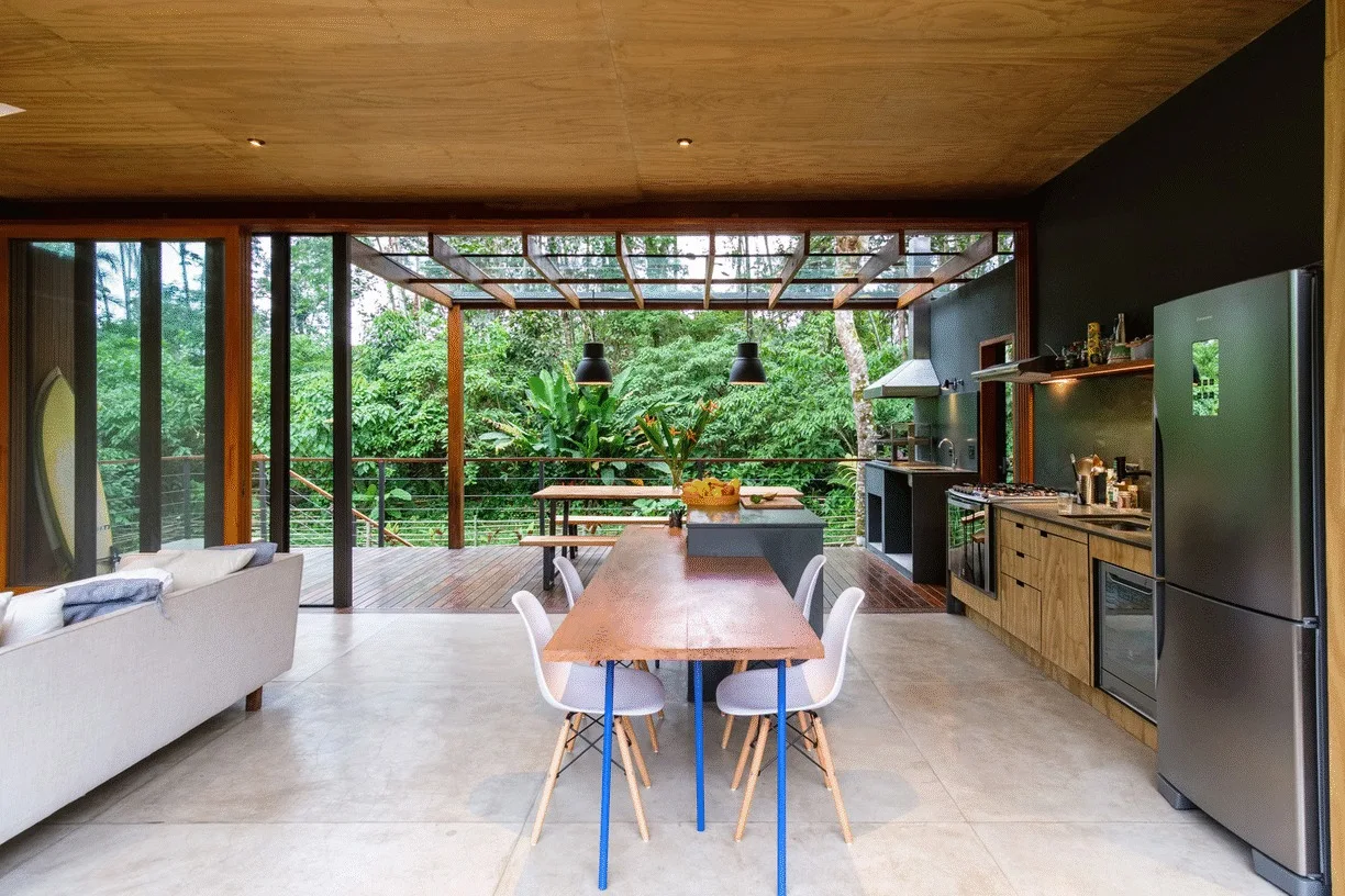 A modern kitchen and dining area features wooden cabinets, a plywood ceiling, and glass doors opening to a lush jungle terrace.
