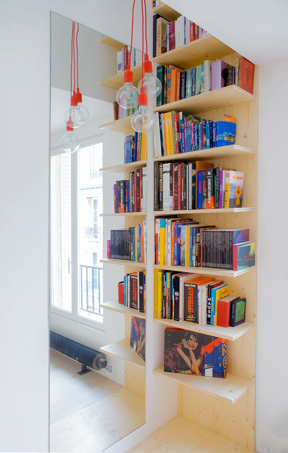 A tall light wood open bookshelf stands next to a vertical mirror that reflects a window and hanging red pendant light cords.