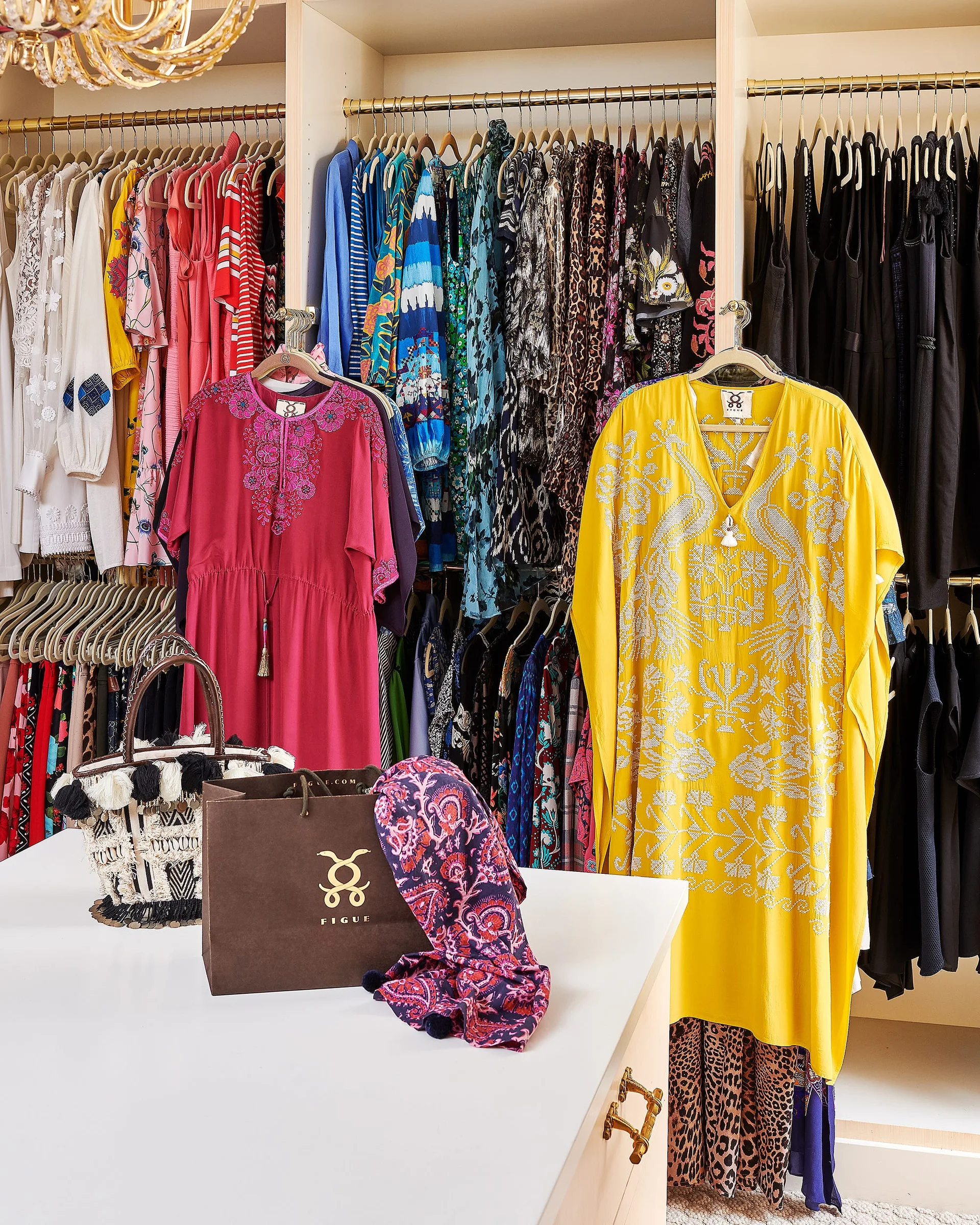 A walk-in closet features built-in white shelves with gold hanging rods filled with colorful clothing, and a white island displaying bags and a scarf.