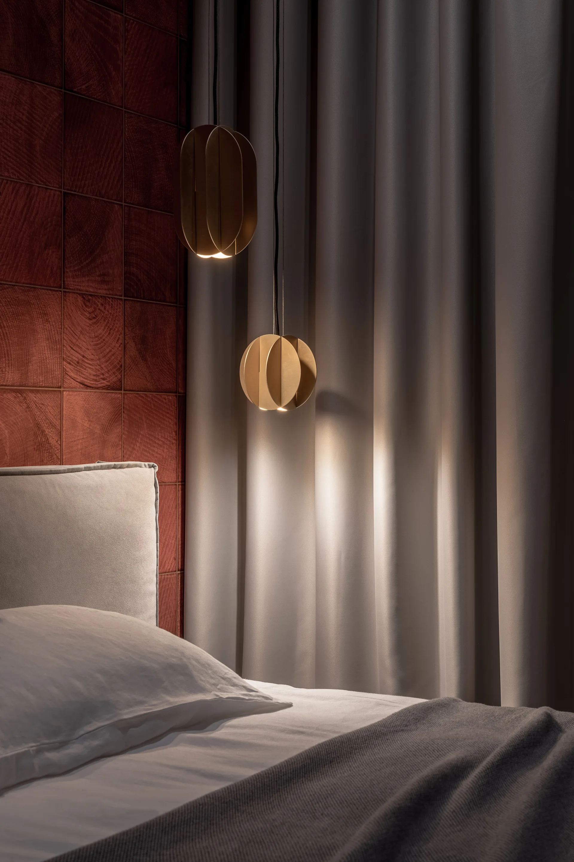 A bedroom features a bed with a grey headboard, a dark red grid wood paneled wall, and two staggered bronze pendant lights against grey drapery.