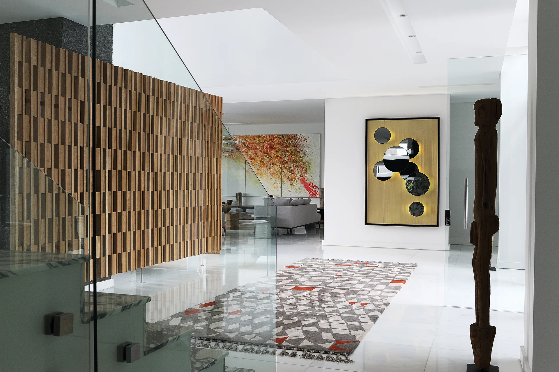 A modern foyer features a slatted wood partition, a glass staircase railing, a geometric patterned rug, and a backlit circular wall art piece.