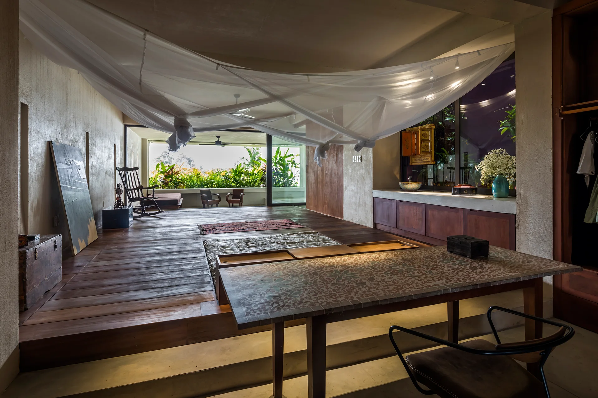 A rustic bedroom features a raised wooden platform with a bed under a mosquito net canopy, a mosaic table, and a window to a lush terrace.
