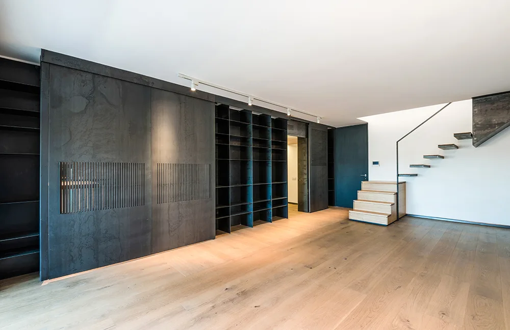A minimalist room features a large matte black sliding storage wall alongside an open riser wooden staircase on light hardwood floors.