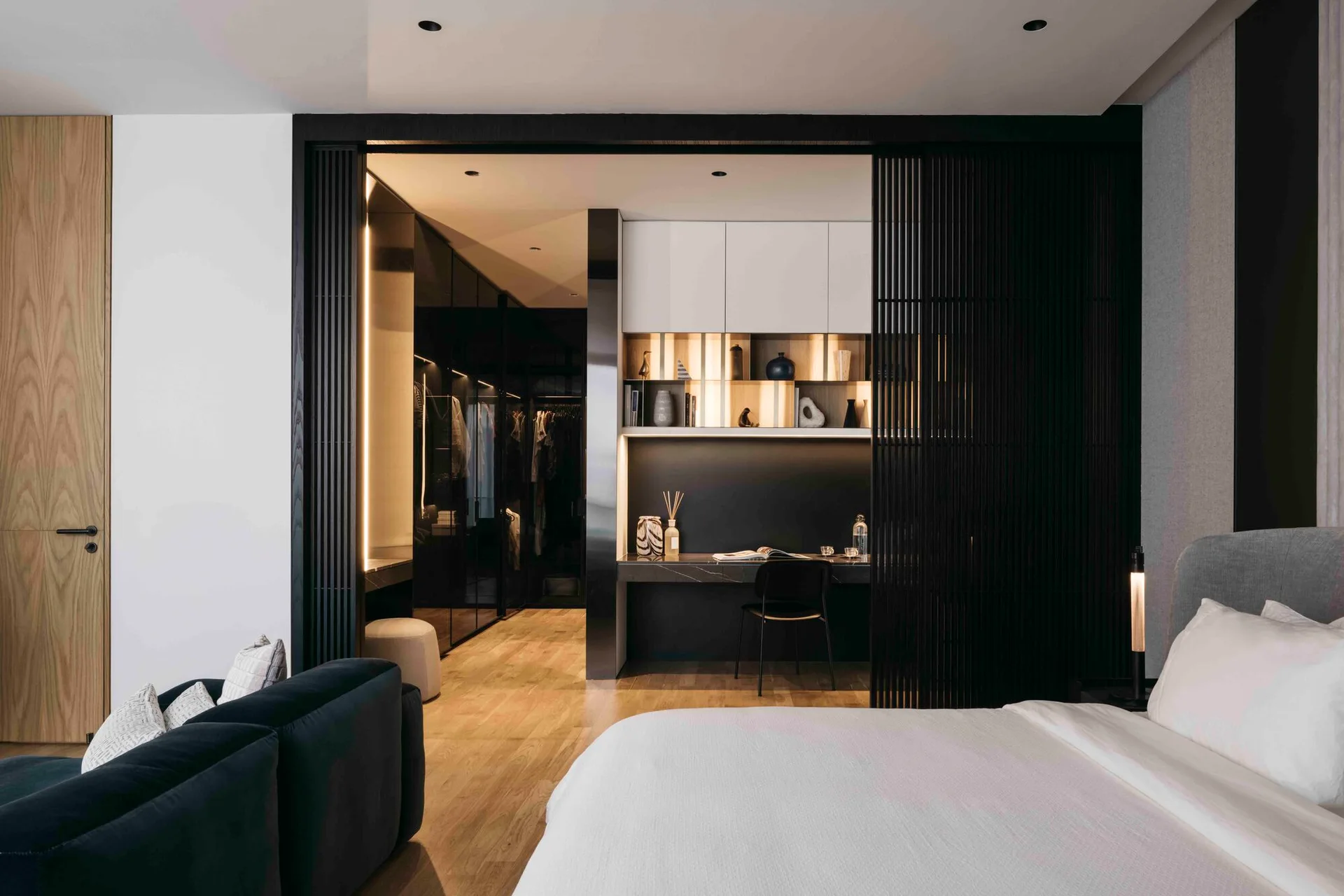A modern bedroom features a white bed, a dark slatted sliding door, and an integrated desk alcove leading to a walk-in closet.
