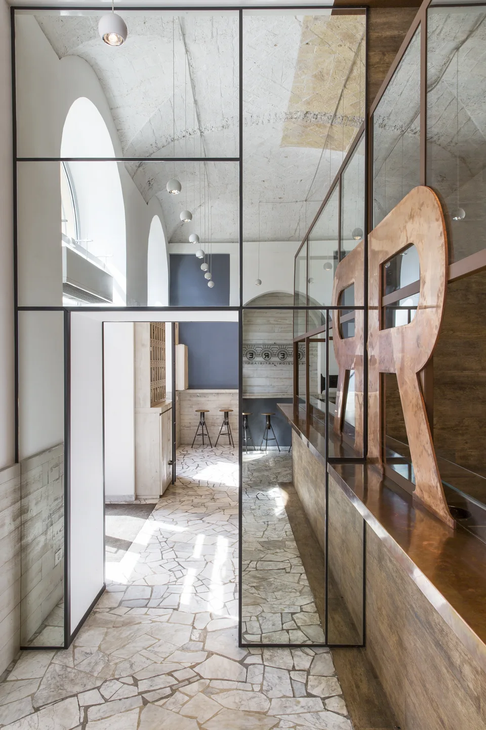 An interior view of a commercial space with a metal-framed glass partition, irregular stone tile flooring, and a vaulted masonry ceiling.