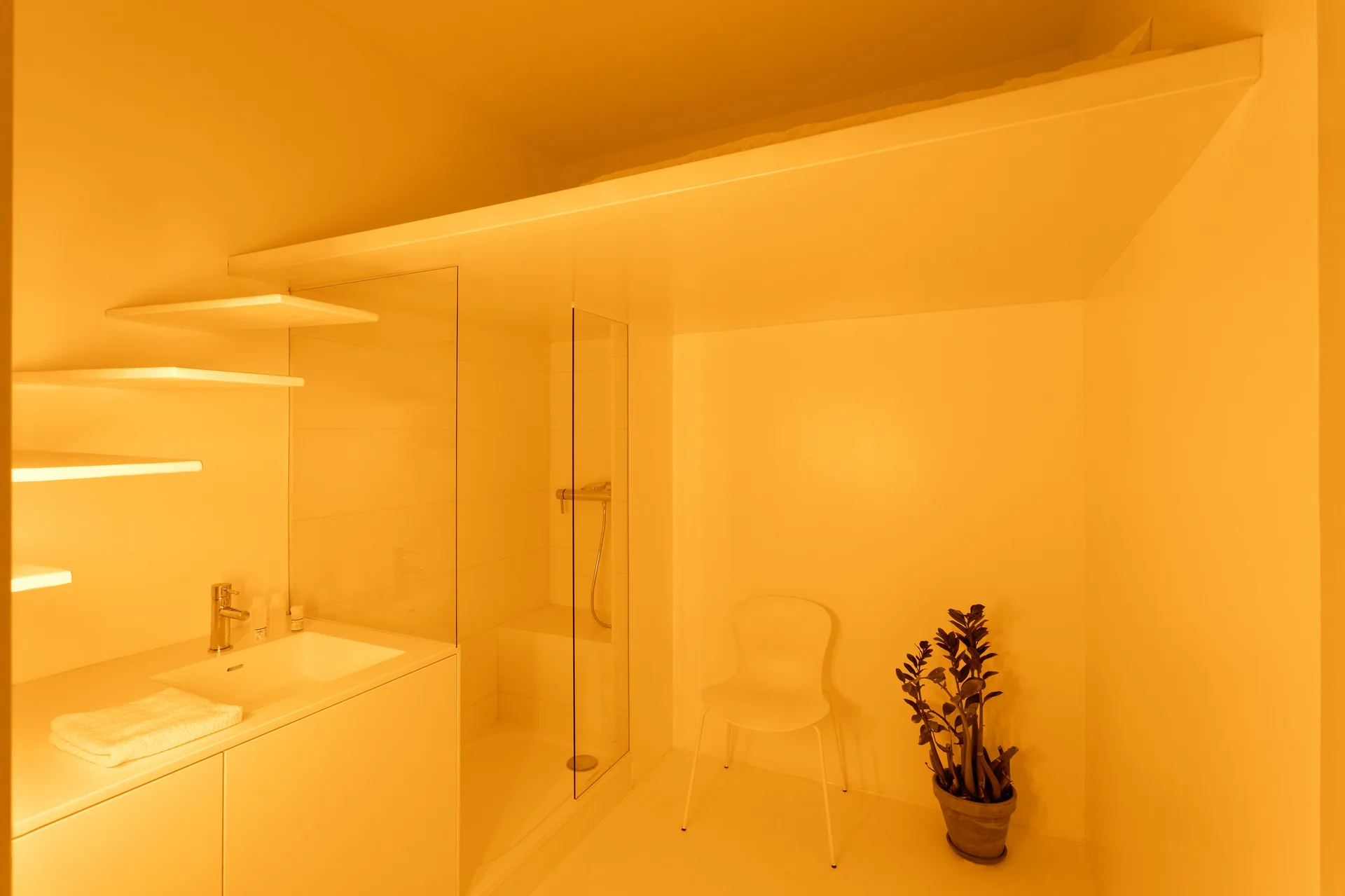 A minimalist white bathroom features a seamless vanity, vertical floating shelves, a glass walk-in shower, a simple white chair, and a potted plant.