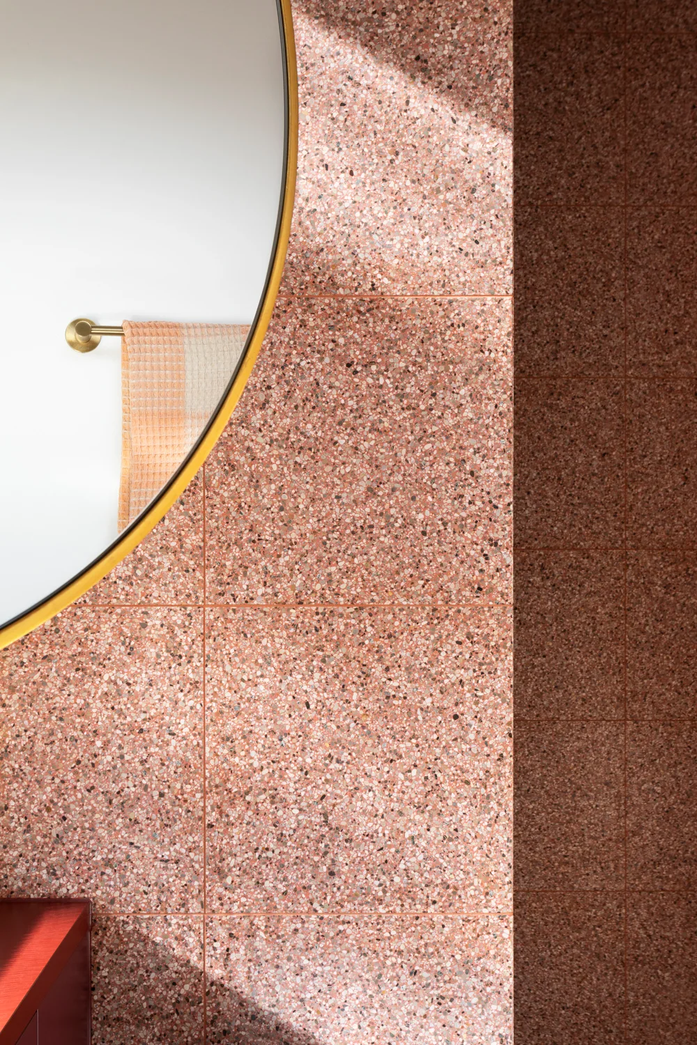 A close-up of a bathroom wall featuring square pink terrazzo tiles and a large circular mirror reflecting a peach-colored towel.