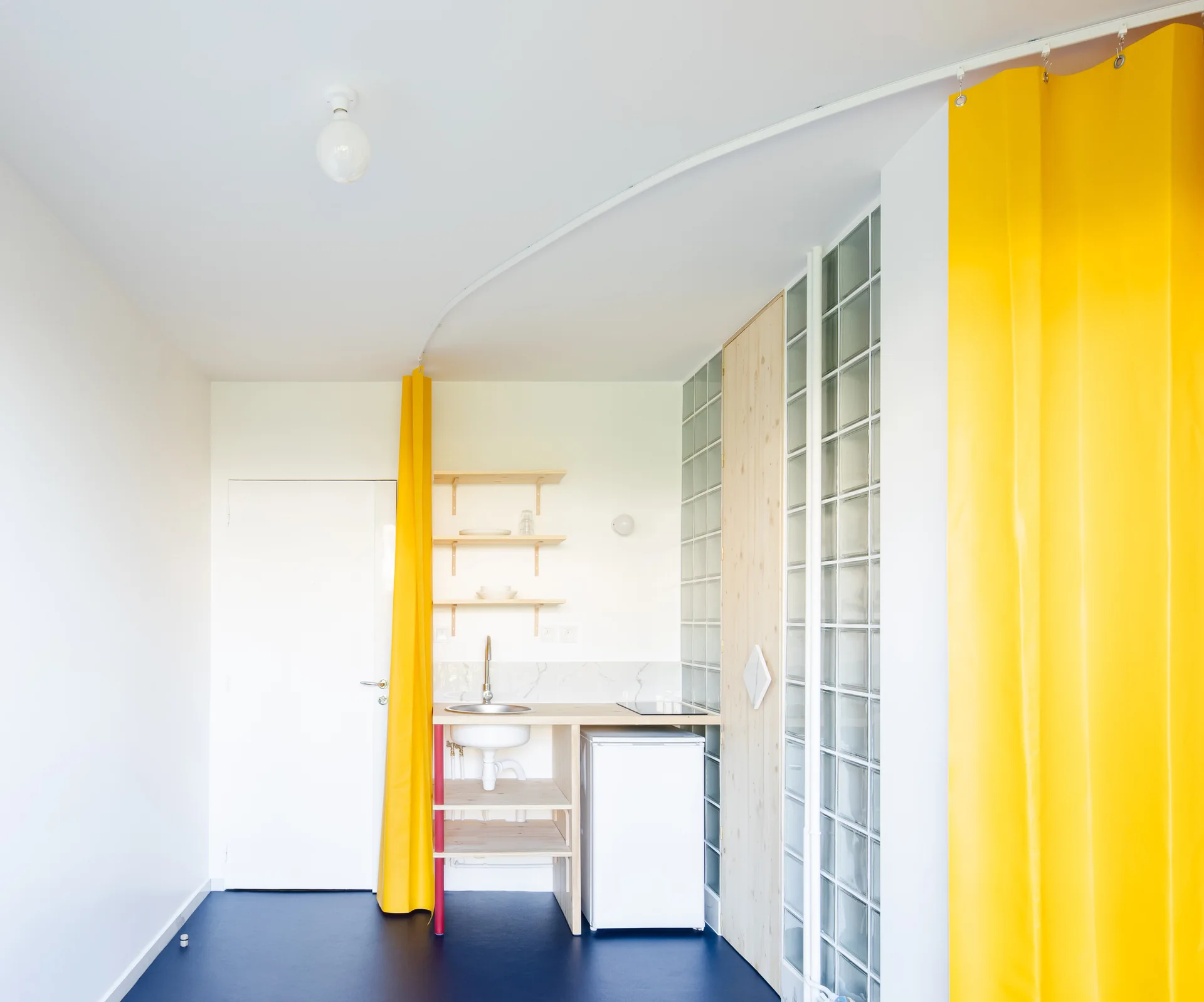 A minimalist kitchenette with wood shelving, a glass block wall, and a bright yellow curtain hanging from a curved track on the ceiling.