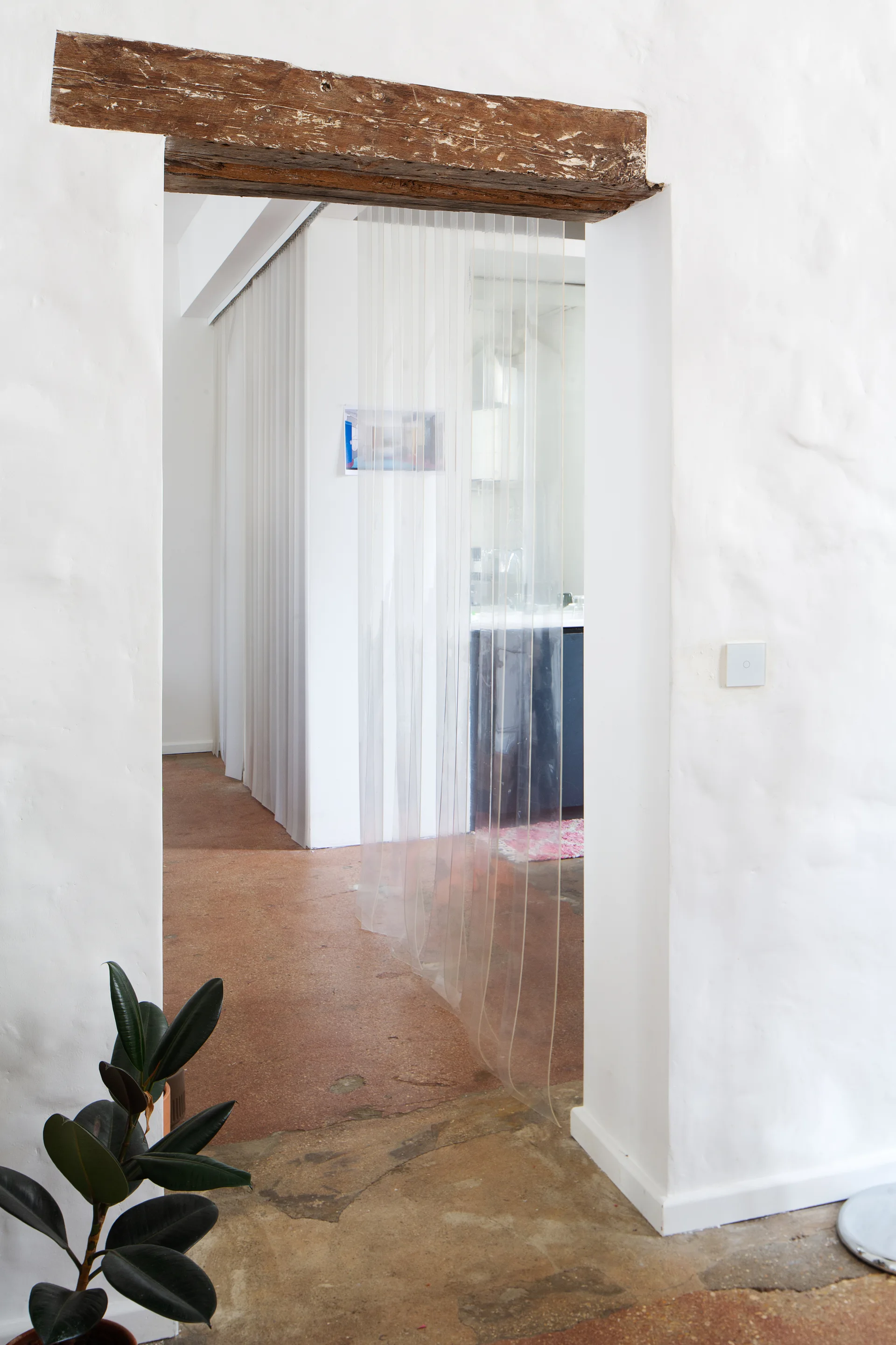 A doorway framed by a dark rustic wood beam and textured white plaster walls features a clear vinyl strip curtain hanging in the opening.