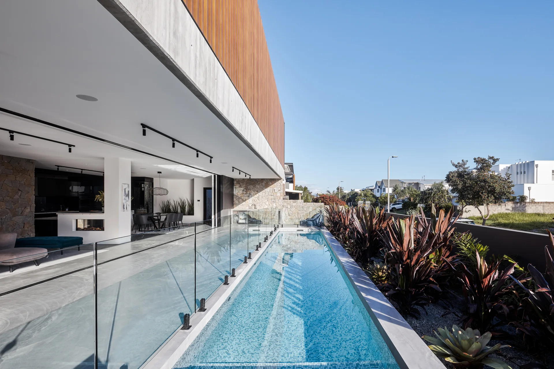 A narrow blue lap pool runs alongside a covered polished concrete patio with a frameless glass railing.