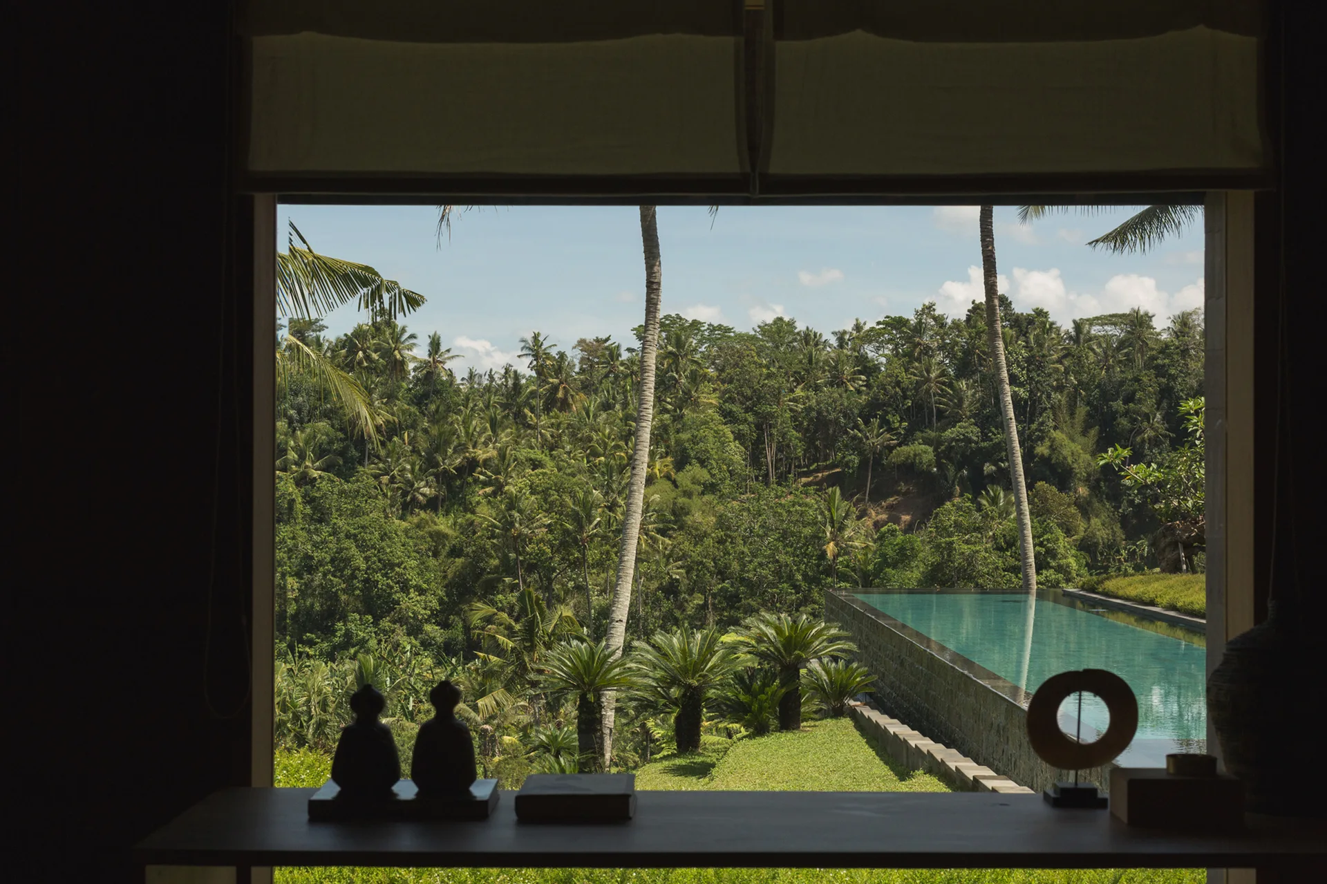 A dark indoor console table with sculptural decor sits before a large window looking out at a tropical forest and a turquoise infinity pool.
