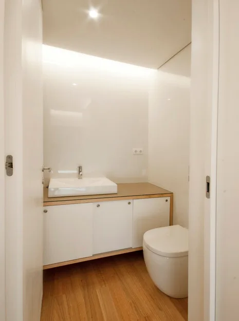 A minimalist powder room seen through a doorway, featuring a white floating vanity, a rectangular washbasin, a modern toilet, and wood flooring.