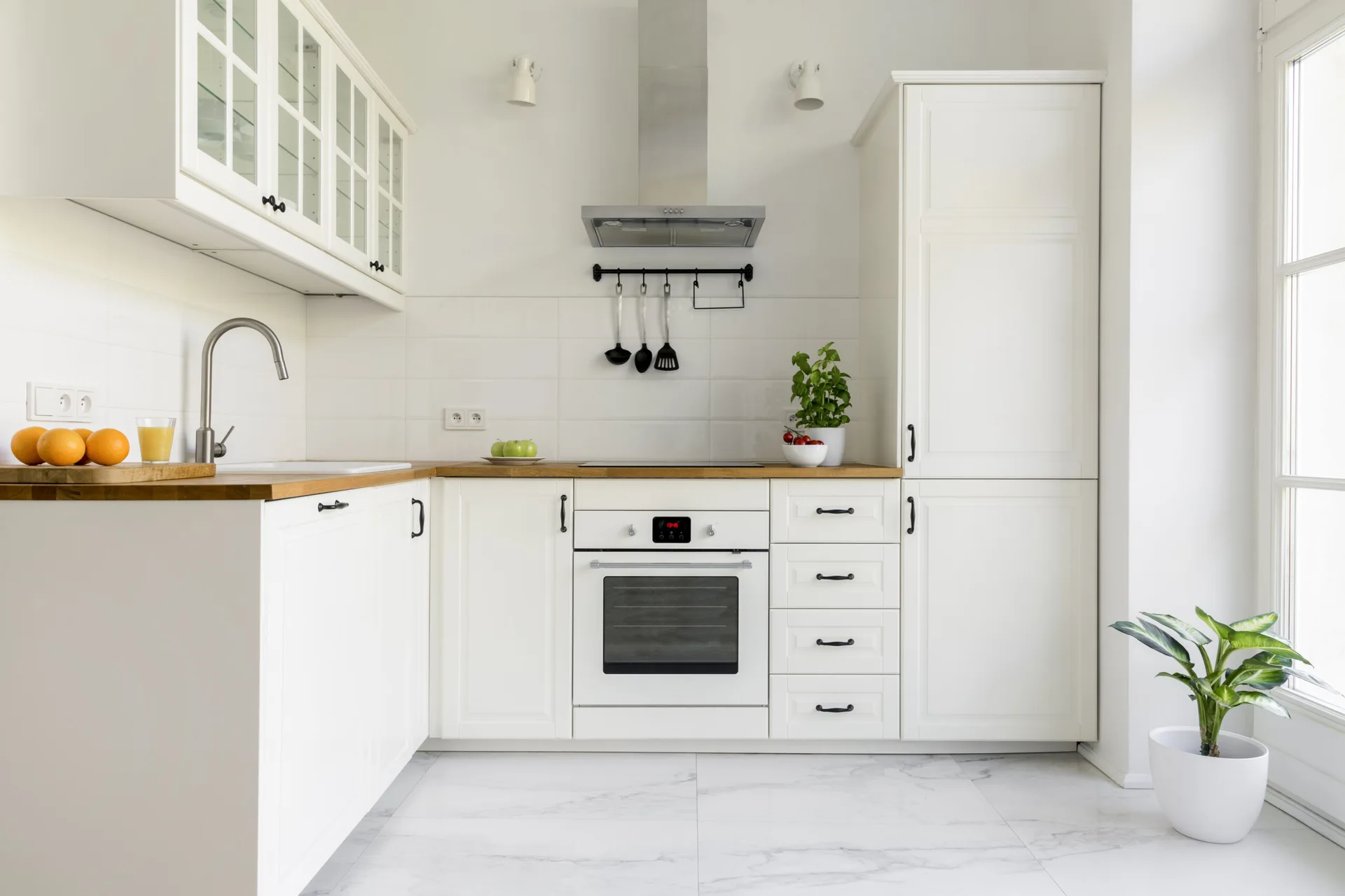 A white L-shaped kitchen featuring white shaker cabinets with black hardware, natural wood countertops, and white marble-look floor tiles.