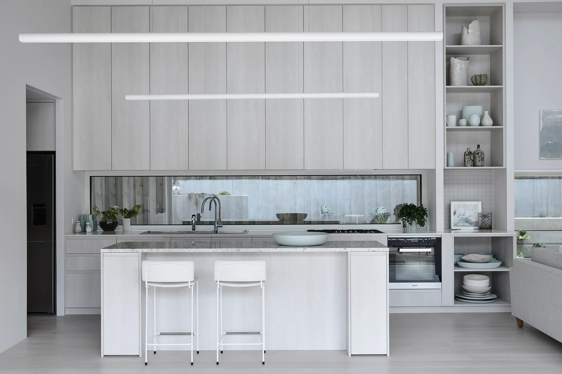 A minimalist kitchen features light wood cabinets, a horizontal ribbon window, a central island with two white bar stools, and open display shelves.