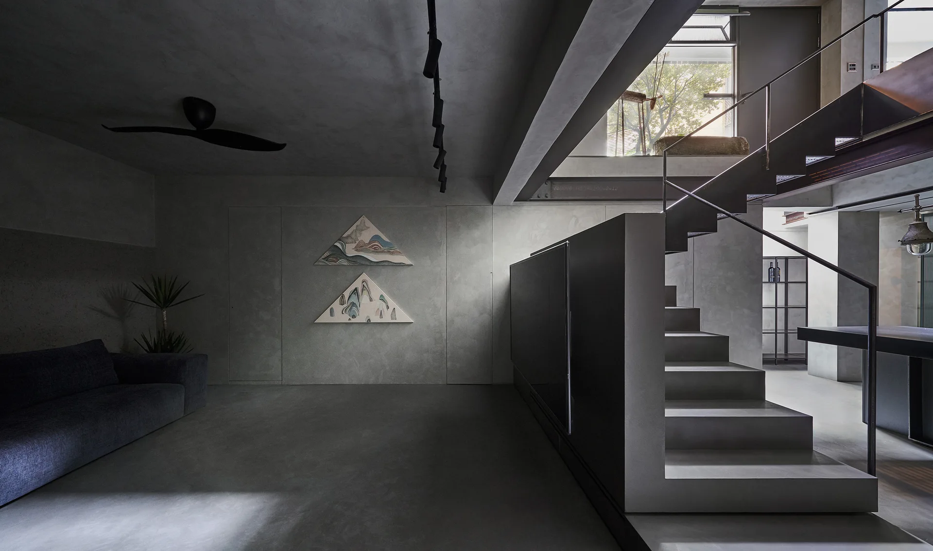 A minimalist concrete living room features a dark low-profile sofa, a central staircase with an integrated media wall, and triangular wall art.