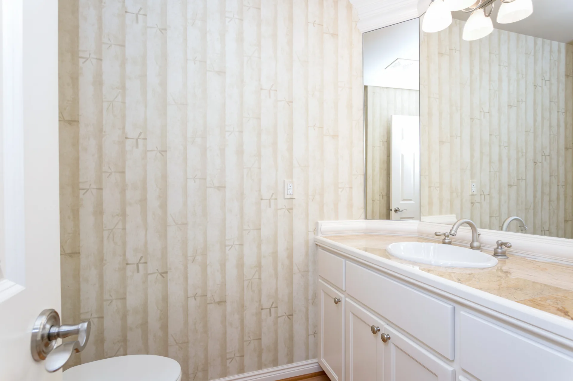 A powder room features a white vanity cabinet with a beige stone countertop, a large mirror, and bamboo-patterned wallpaper.