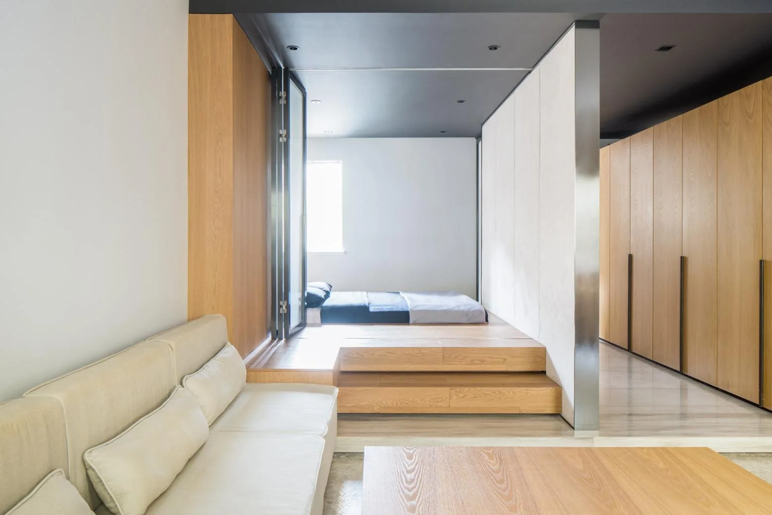 A modern minimalist studio space features a beige sofa in the foreground and a bedroom mattress resting on an elevated wooden platform with steps.