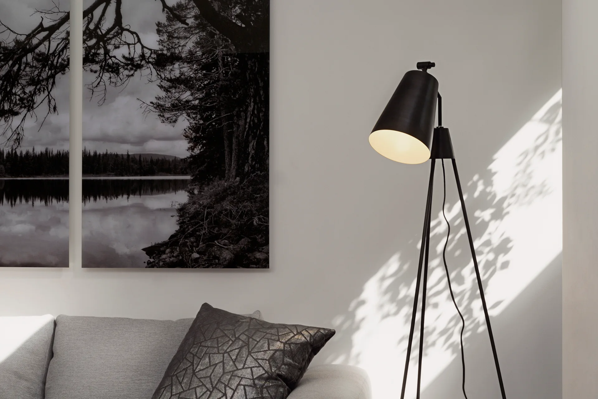 A light grey sofa with a dark patterned throw pillow sits next to a black tripod floor lamp and black-and-white landscape wall art.