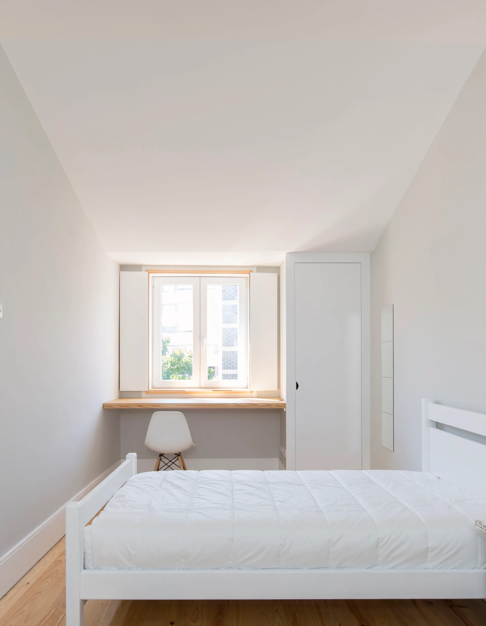A minimalist white bedroom with a white bed, a built-in light wood desk underneath a window, and folding white shutters.