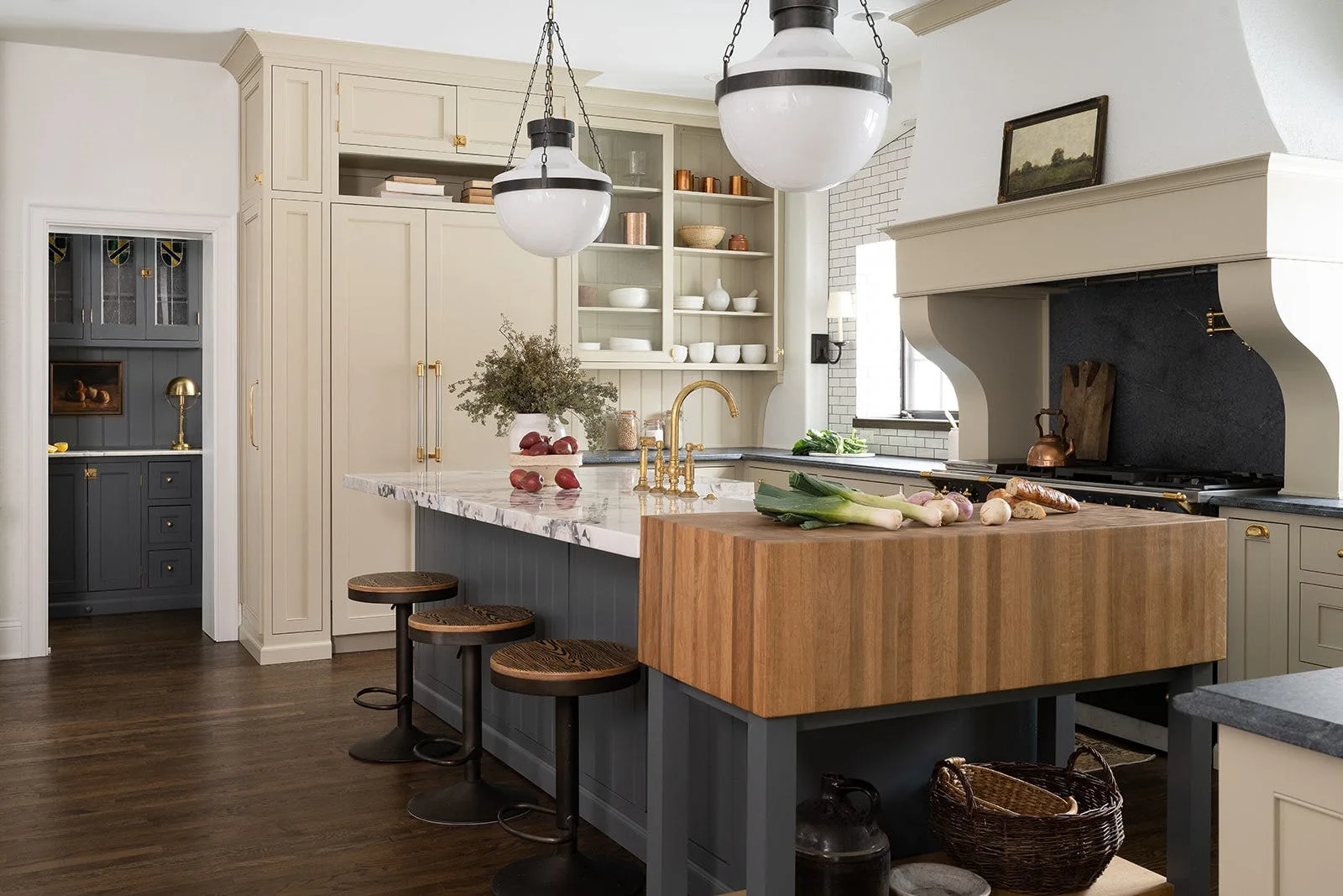 A kitchen features a dark grey island with a marble top and a thick wood butcher block extension, surrounded by beige cabinets and round bar stools.