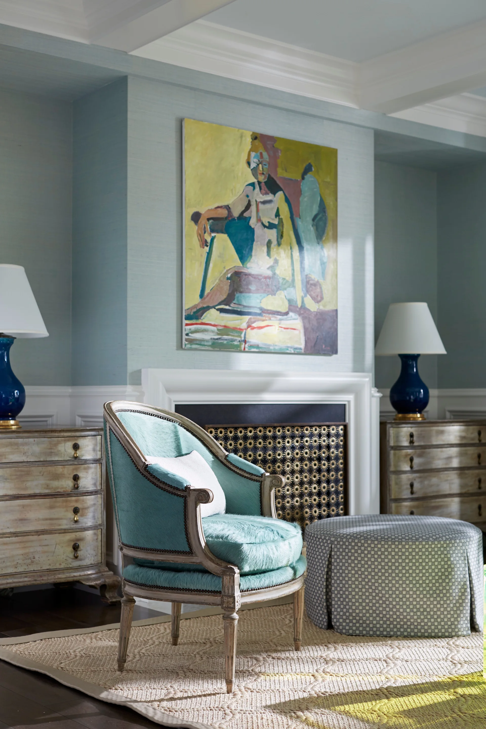 A traditional sitting area featuring a teal armchair, patterned ottoman, and two distressed wood chests flanking a fireplace with a large painting.