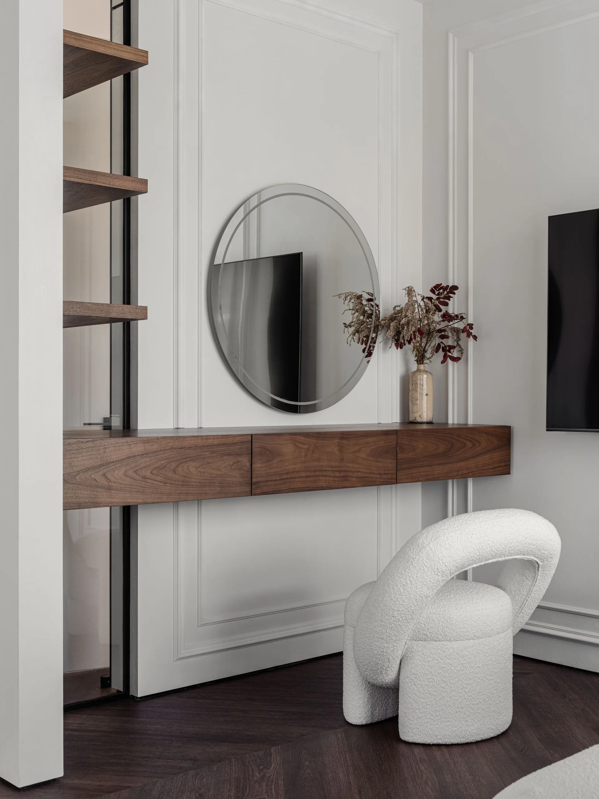 A modern bedroom vanity area featuring a floating wooden console, a circular wall mirror, and a white sculptural boucle chair against paneled walls.