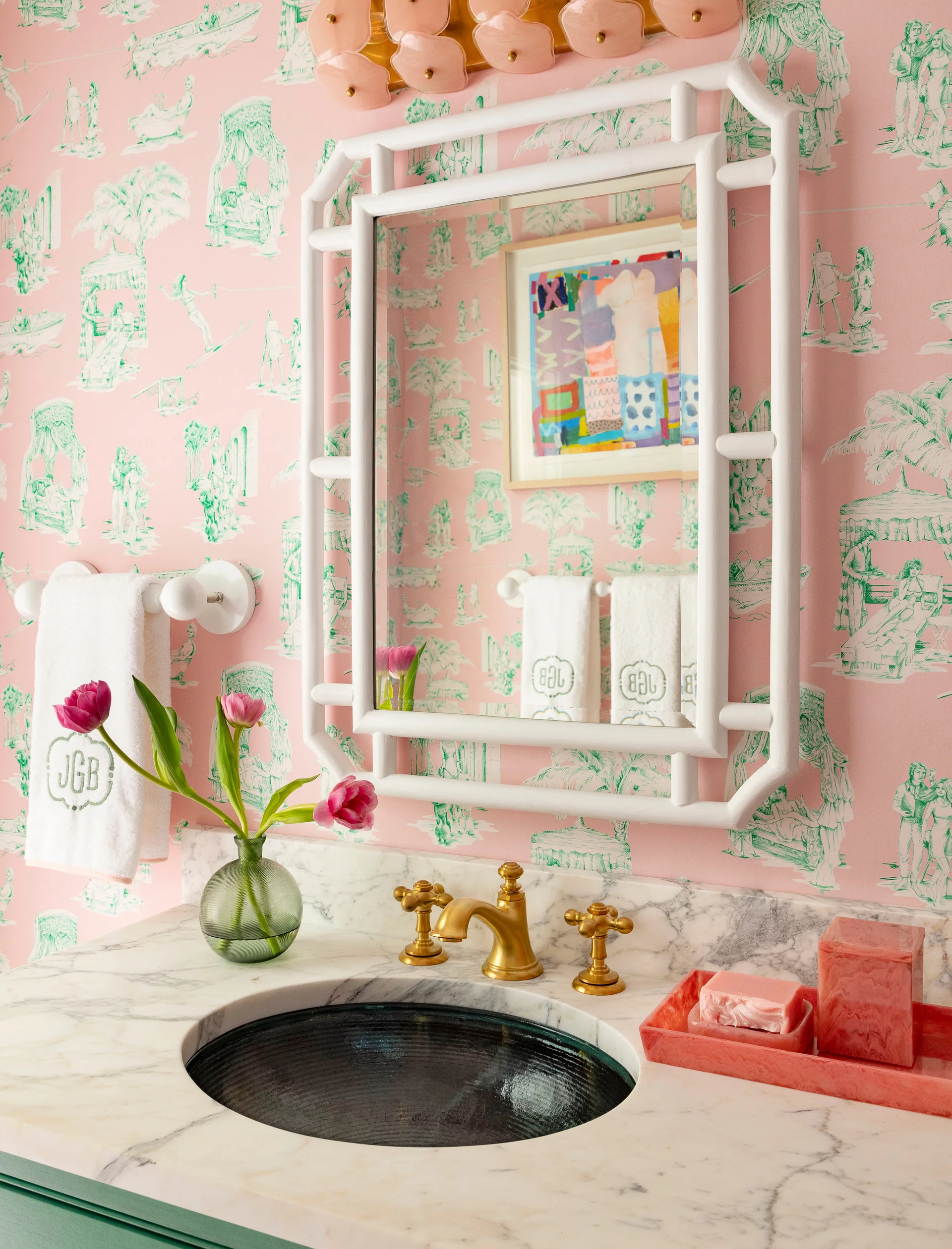 A bathroom vanity features a white marble countertop, a brass faucet, and a white bamboo-framed mirror against pink and green toile wallpaper.
